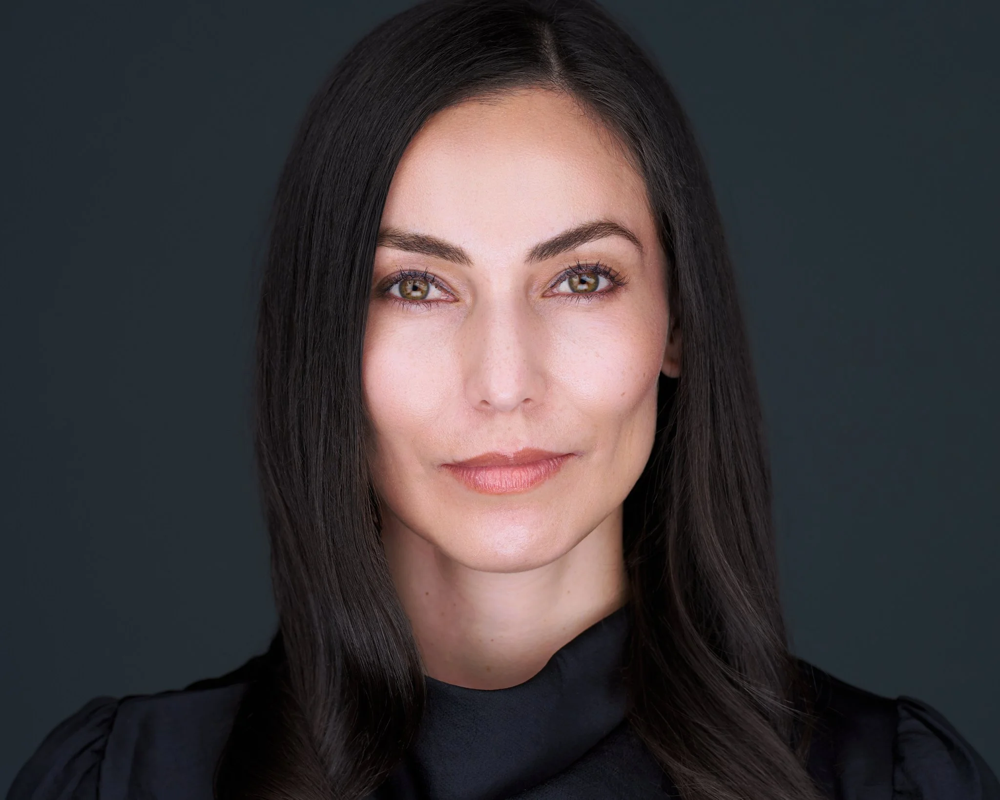 Close-up of a woman with straight black hair, light skin, and brown eyes, wearing a black top, against a dark background.