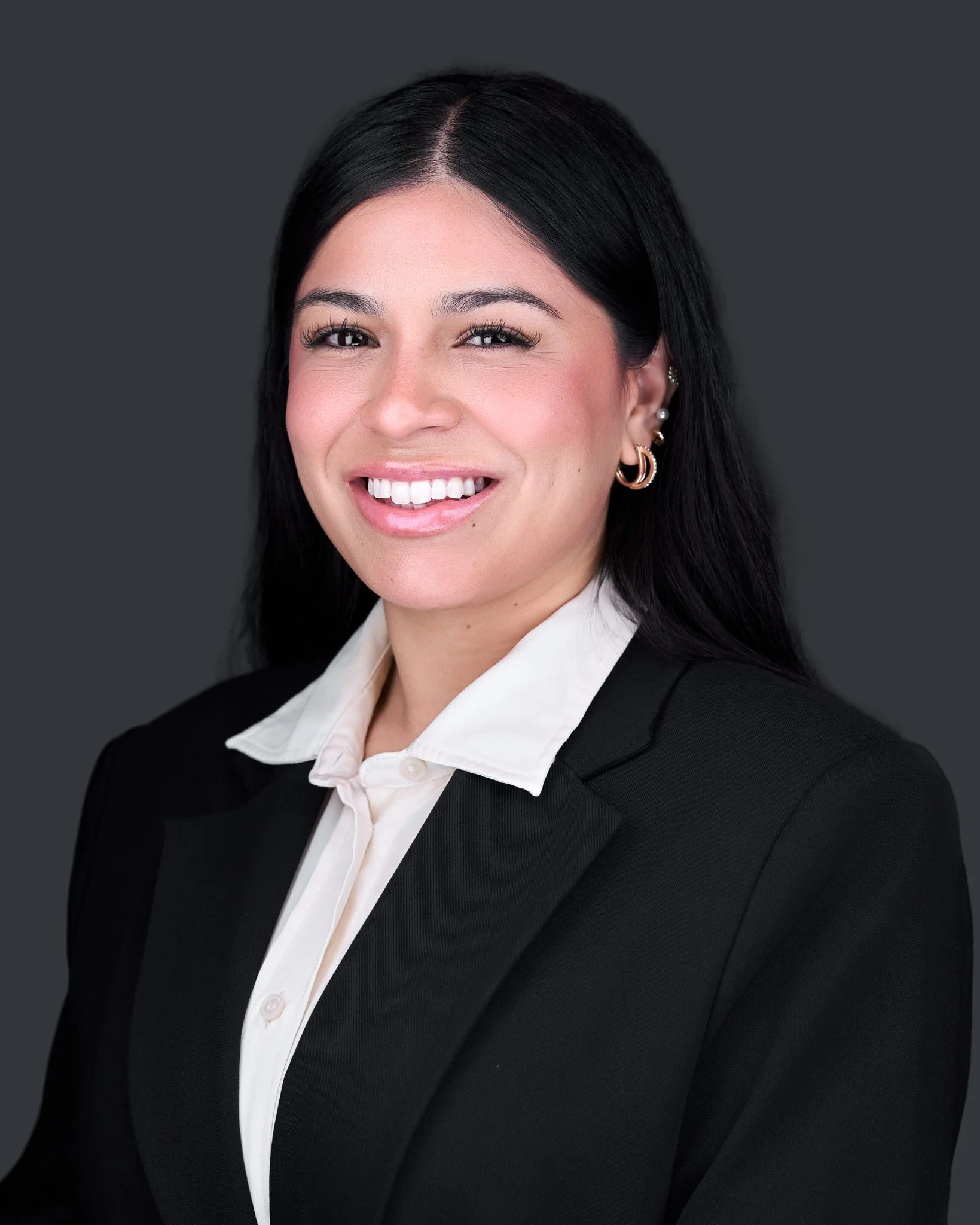 A woman with long black hair, wearing a black blazer over a white collared shirt, smiling and looking at the camera, with earrings, against a solid dark gray background.