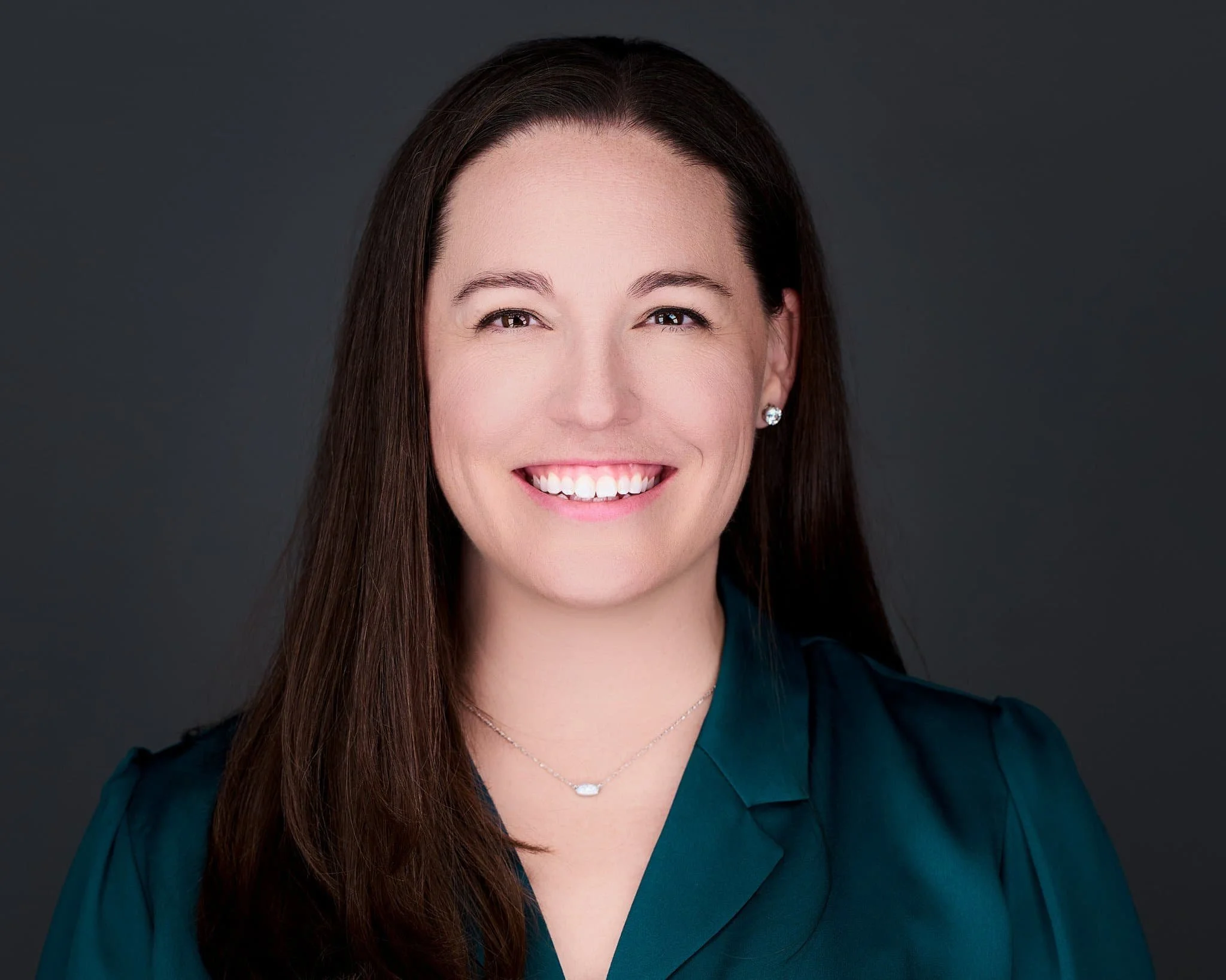 A woman with long brown hair smiling, wearing a teal blouse, earrings, a necklace, and standing against a dark gray background.