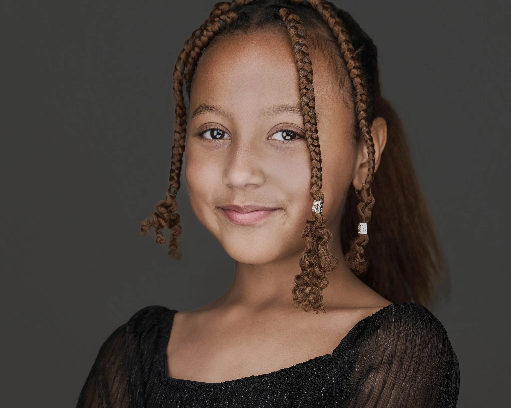 A young girl with brown skin and braided hair with silver beads, wearing a black tulle dress, smiling against a dark grey background.