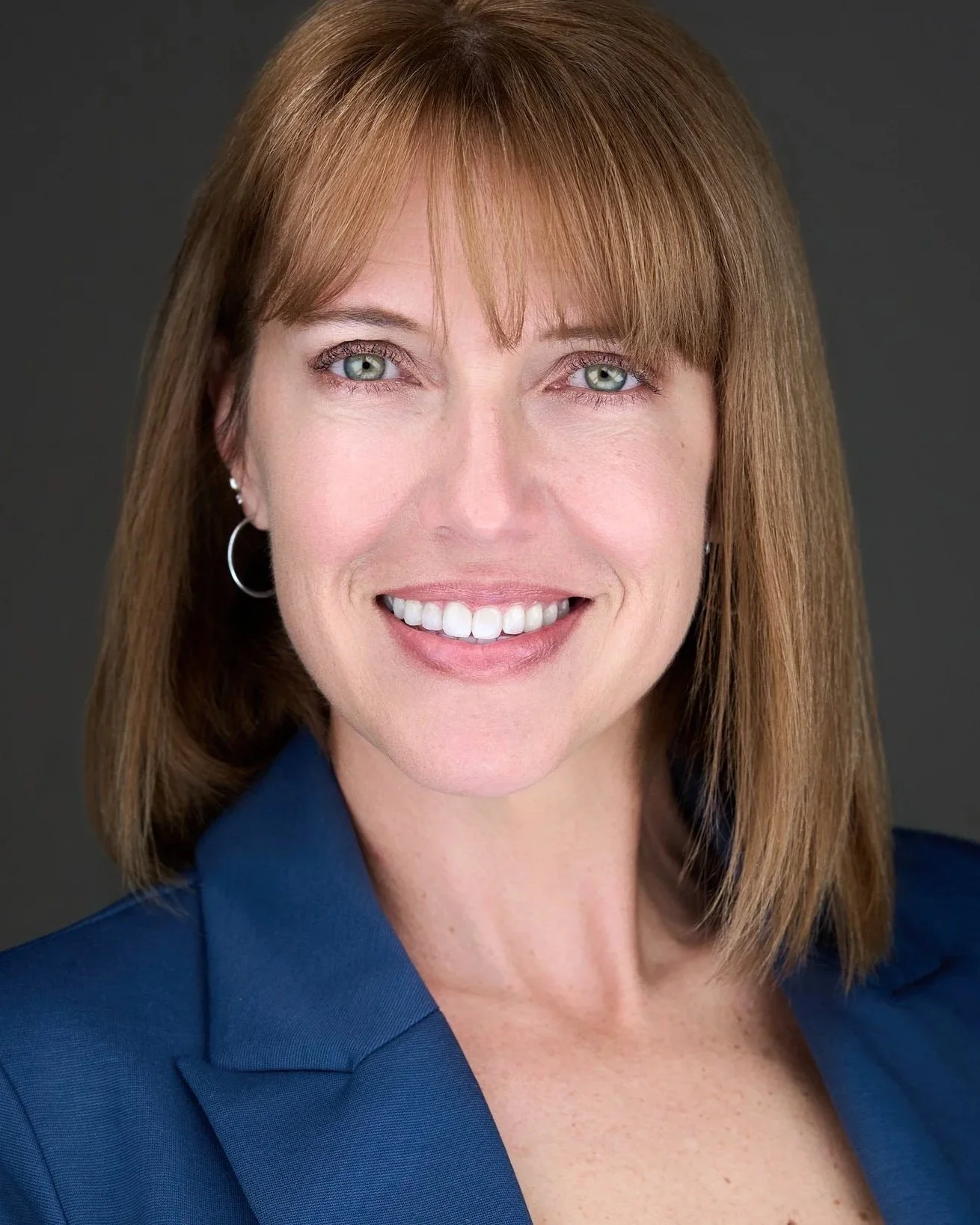 Headshot of a woman with reddish-brown hair, blue eyes, wearing a blue blazer and earrings, smiling against a dark background.
