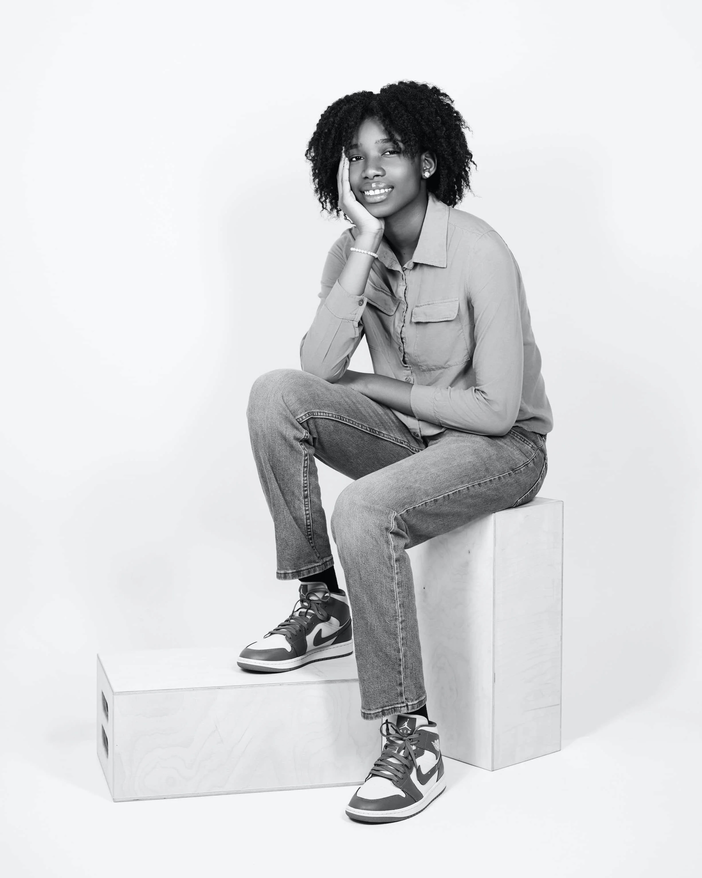 A woman sitting on a wooden box, wearing a button-up shirt, jeans, and Nike sneakers, smiling with her hand resting on her face, against a plain white background.