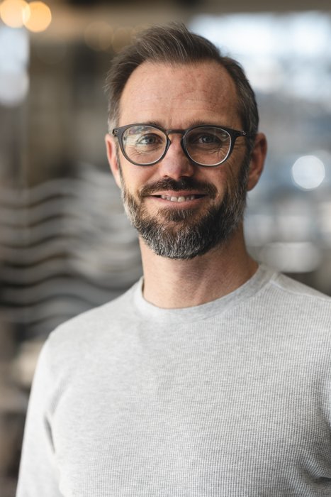 A man with glasses and a beard wearing a black shirt, standing in a modern indoor setting.