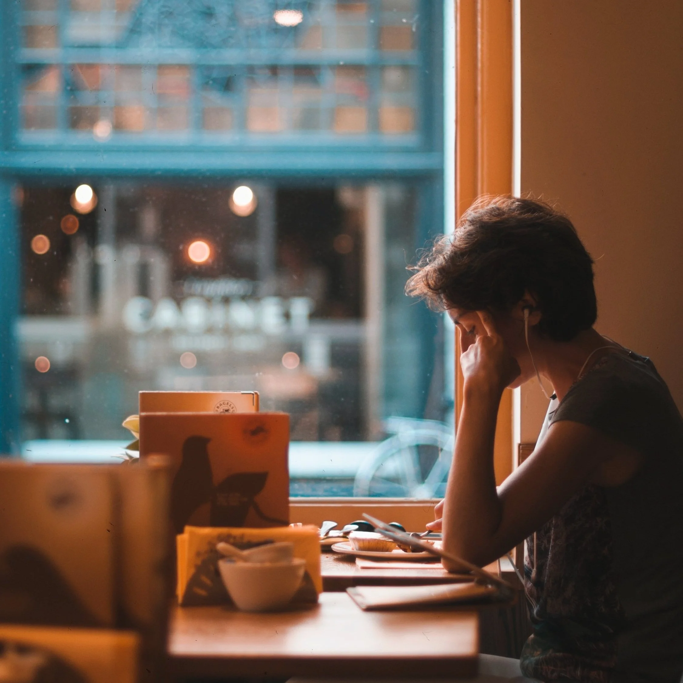 Person sitting at a cafe table next to a window, wearing earphones and looking at a device, with coffee cups and a city street visible outside.