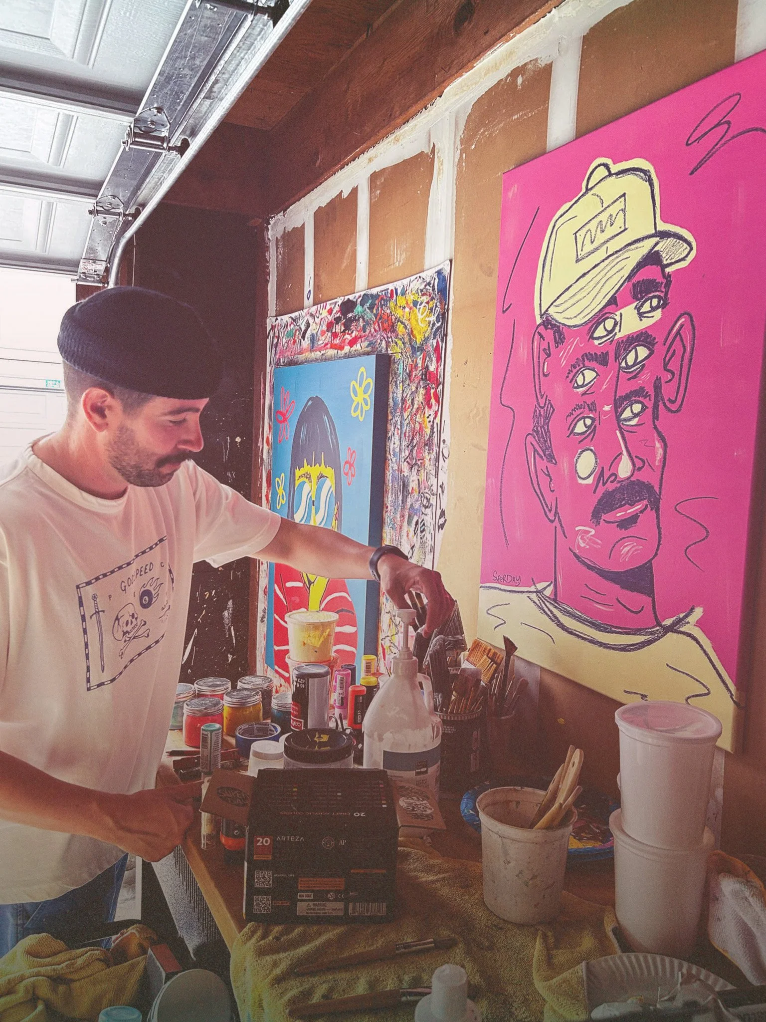 Diego reaching for a brush in his studio. He is wearing a black beanie and white shirt.