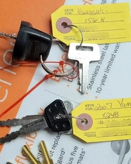 Two sets of car keys with JC Lock & Key tags attached, resting on a paper surface. The keys are for a Kawasaki motorcycle and a Yamaha.