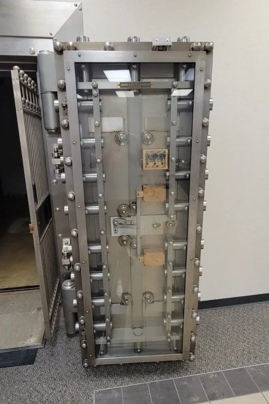 Large, vertical, metal safe vault with numerous bolts and mechanisms, situated indoors on a carpeted floor.