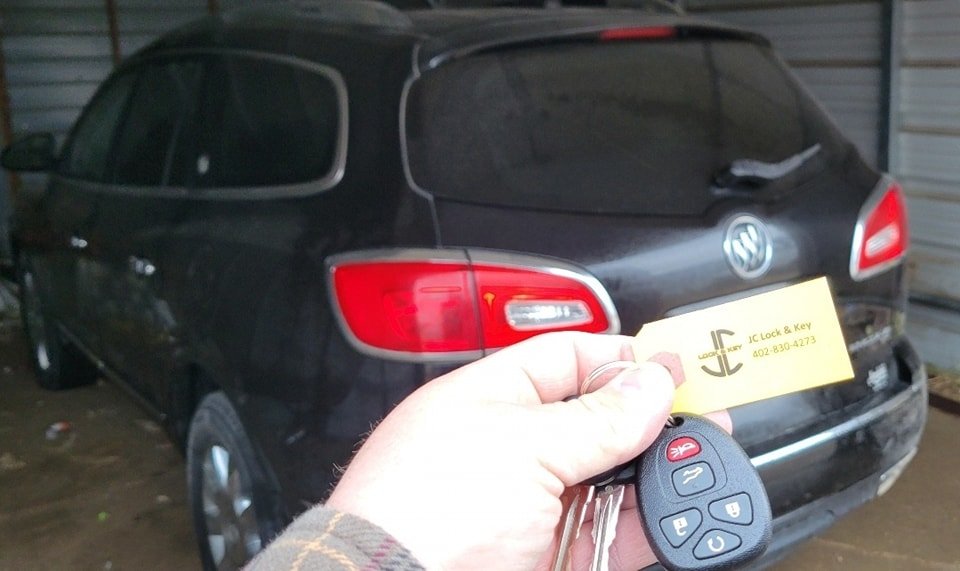 Person holding car keys with a remote key fob, carrying a business card, standing in front of a black Buick SUV in a garage.