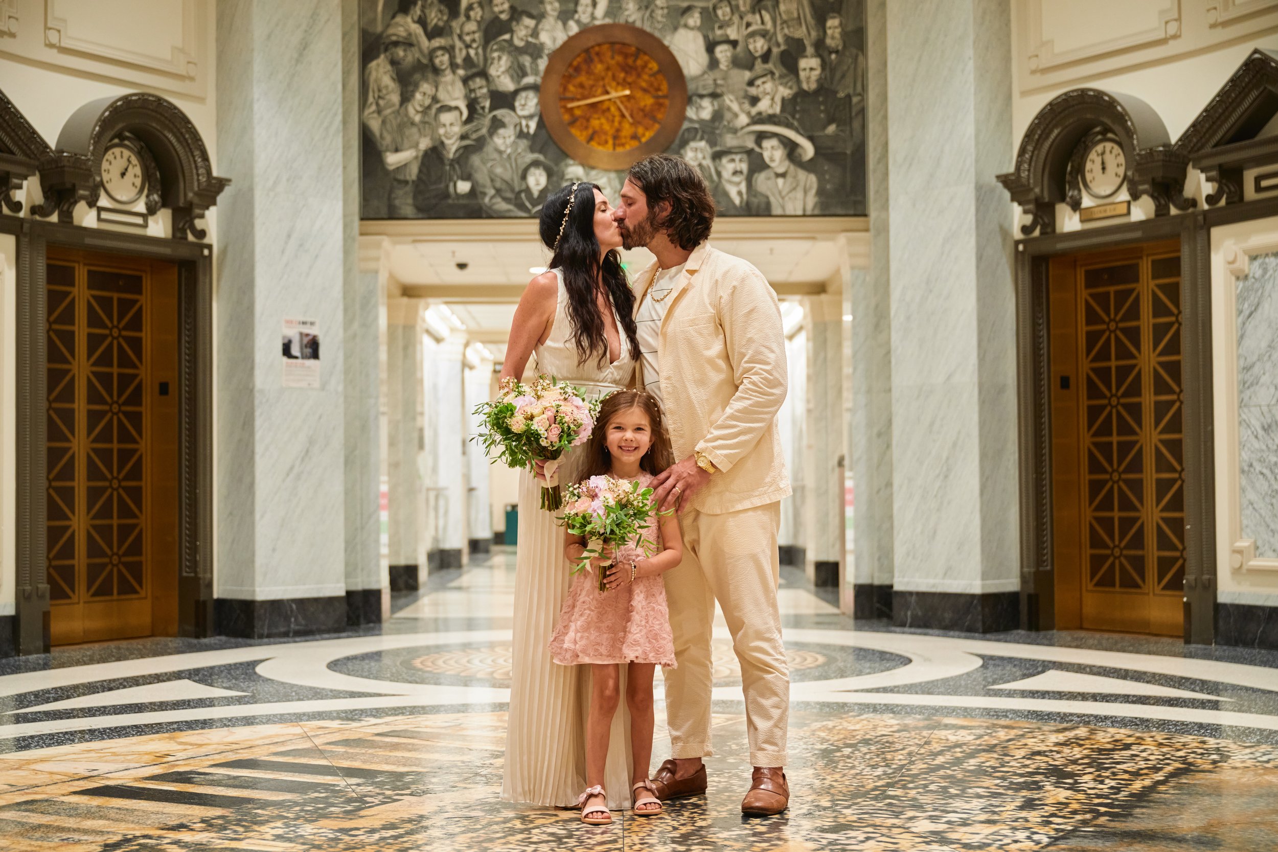 A family of three standing in a historic building, sharing a kiss while holding bouquets, with a mosaic floor and a mural in the background.