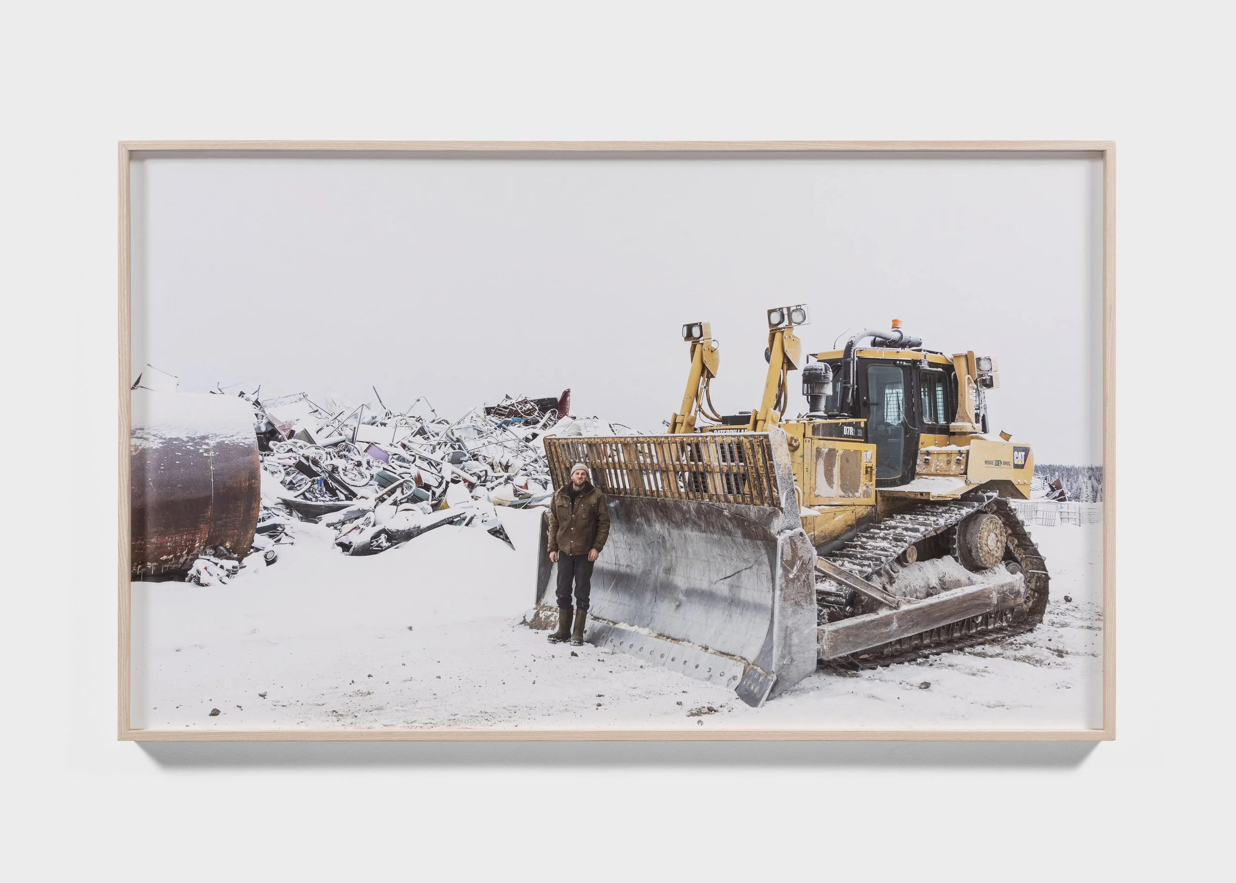 Framed photo of a yellow bulldozer on snow with a person standing next to it, near a pile of scrap metal.