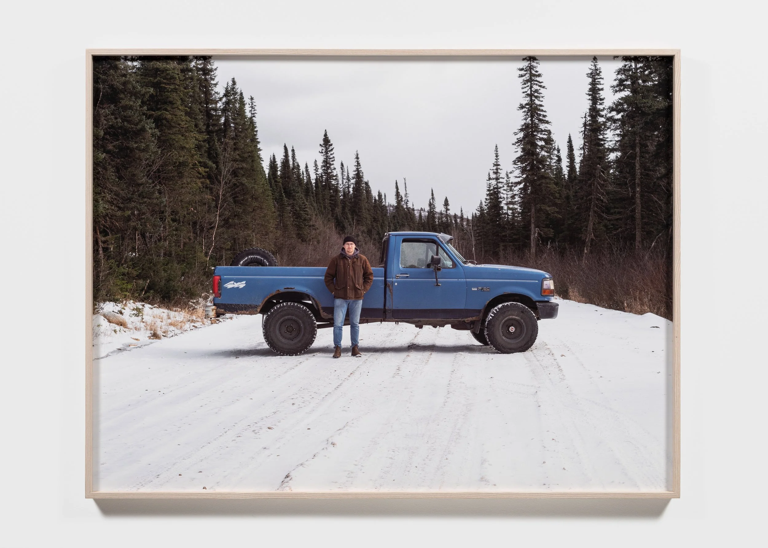 Person standing next to a blue pickup truck on a snowy road surrounded by evergreen trees.