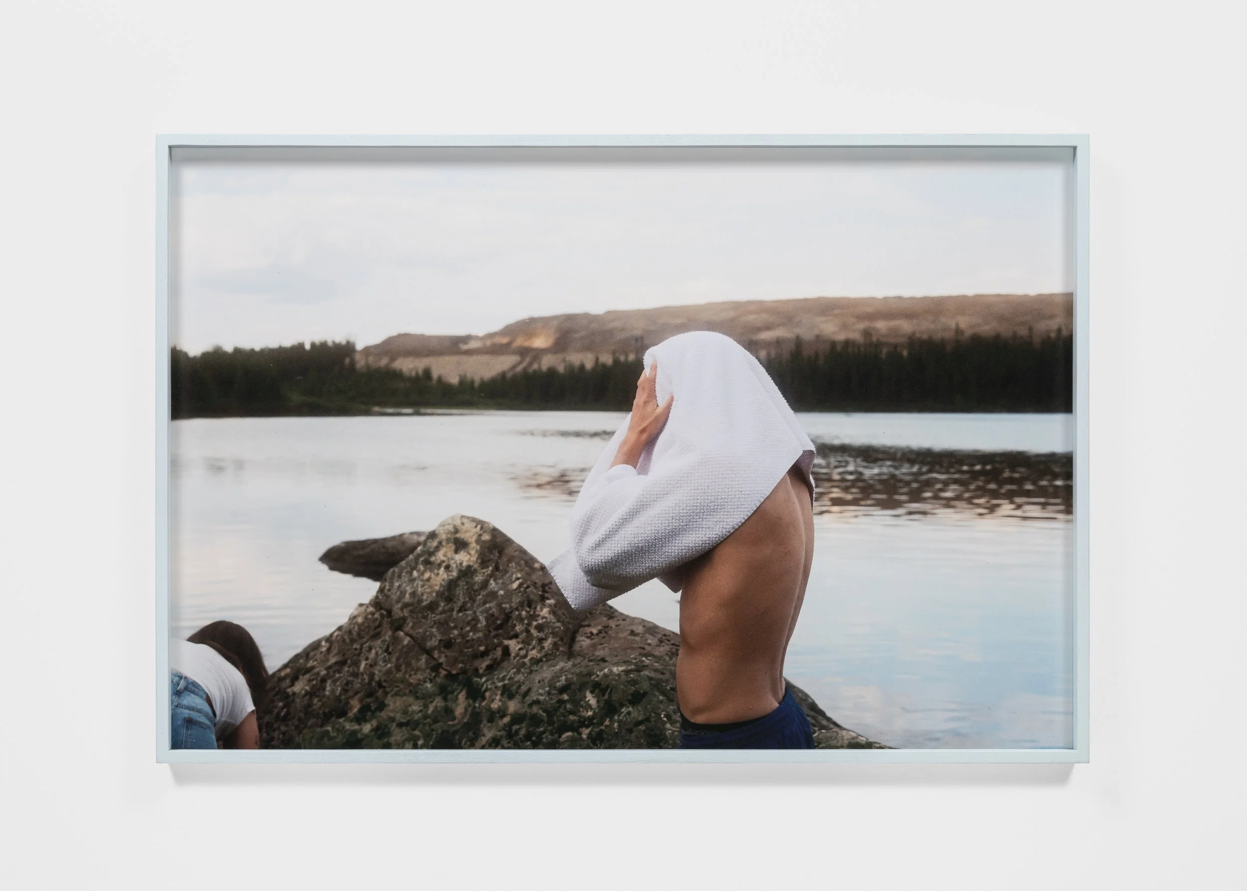 Person drying with a towel by a lake, rocky shoreline, wooded hills in background.