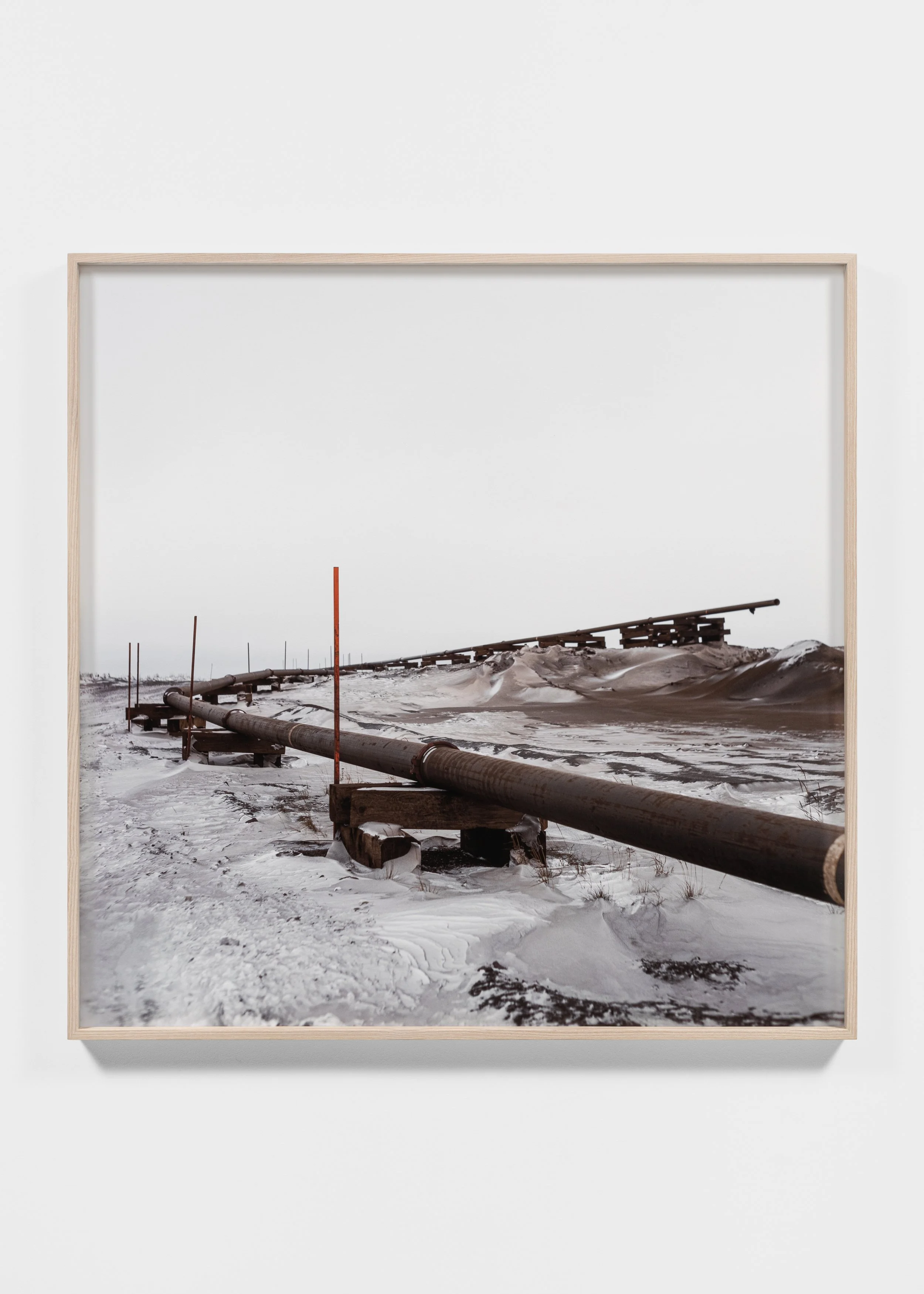 Snow-covered landscape with an elevated pipeline supported by wooden blocks and metal stakes, stretching into the distance.