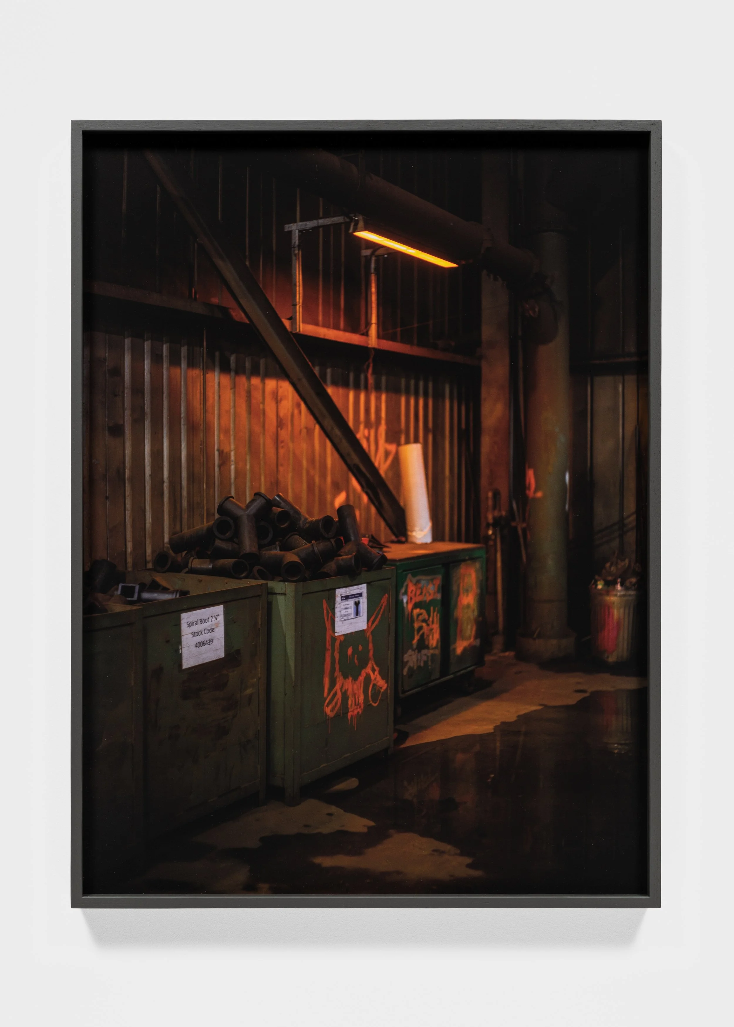 Industrial interior with dim lighting, showing metal and wooden storage containers, graffiti, and scattered metal pipes in a workshop or warehouse setting.