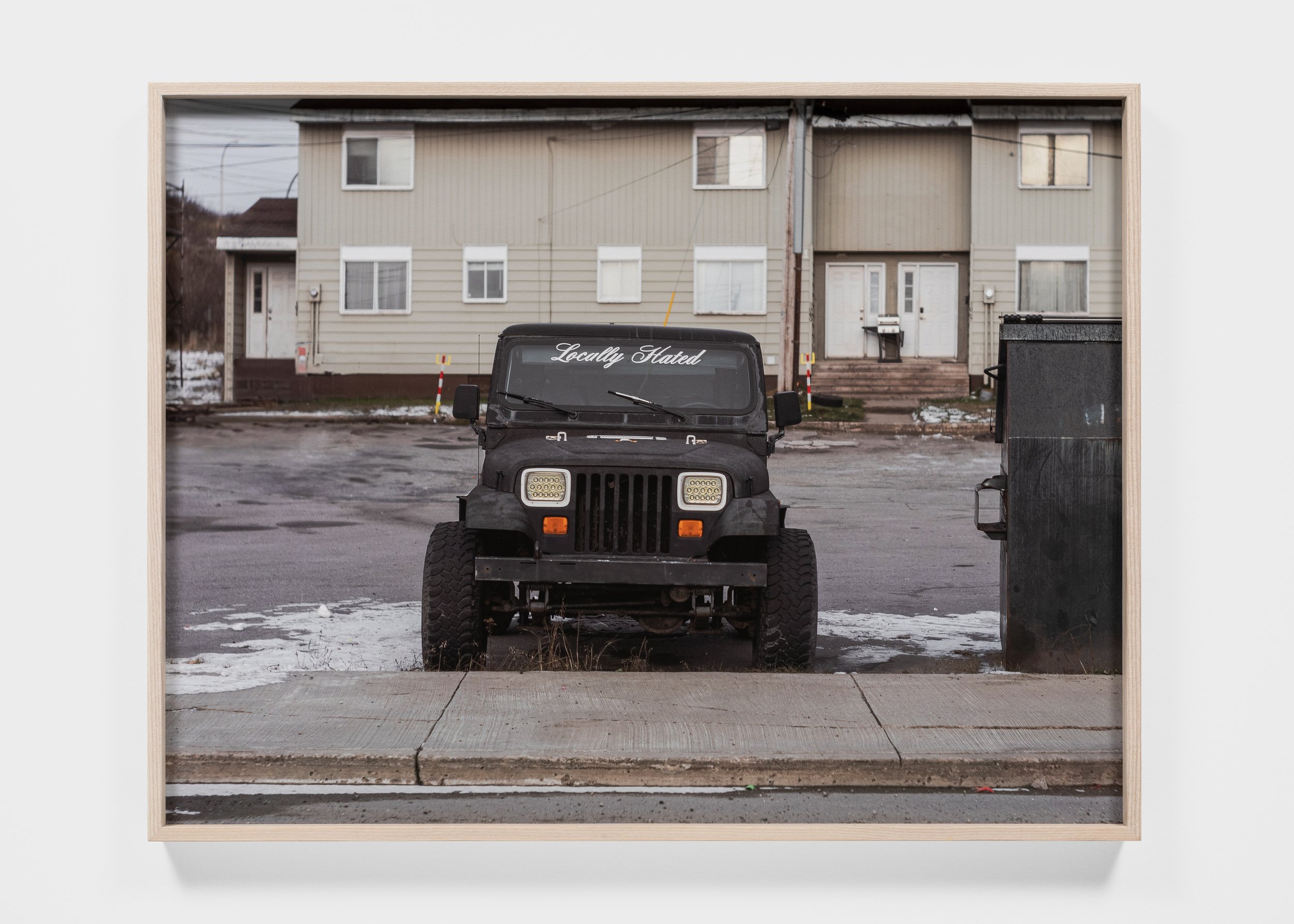 Front view of a parked black Jeep with "Locally Hated" on the windshield, set in front of a beige building with multiple units.