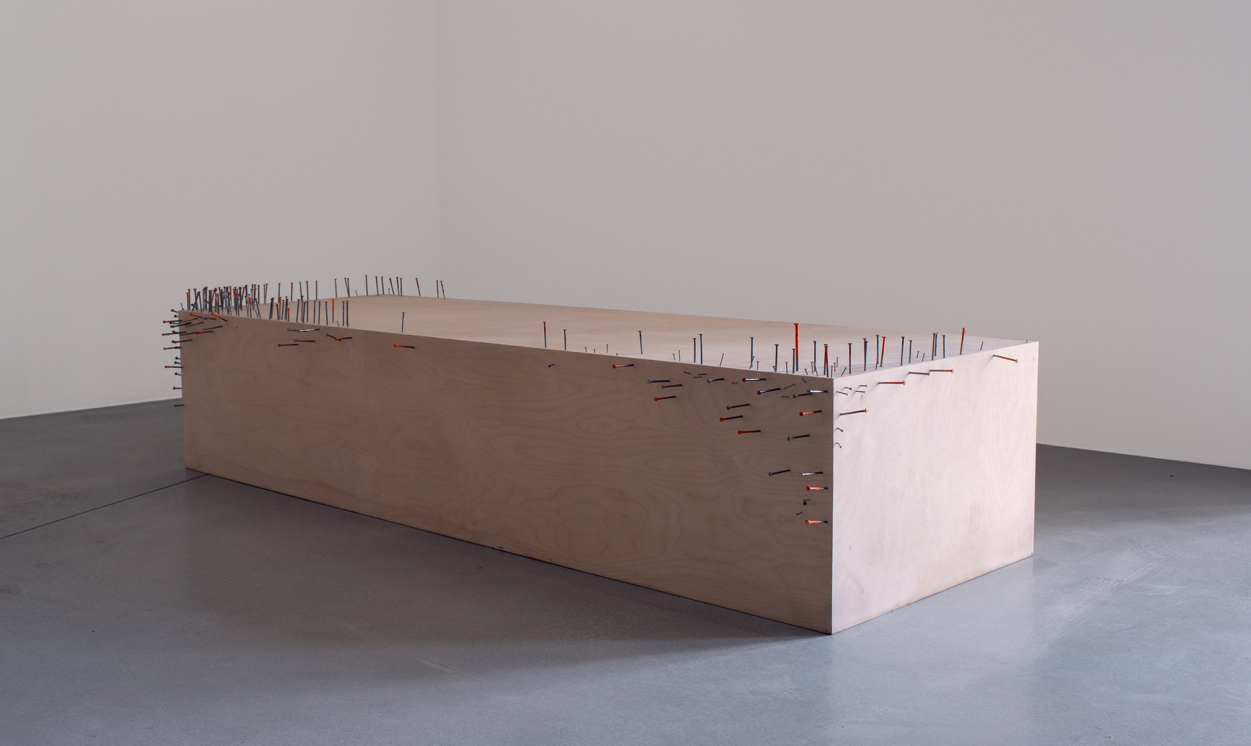 A rectangular wooden block covered with numerous black and red nails protruding from its surface, placed in a minimalist gallery setting on a grey floor.