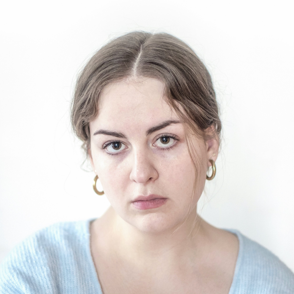 Close-up portrait of a young woman with brown hair, light skin, wearing gold hoop earrings and a light blue top, looking directly at the camera with a neutral expression against a plain white background.