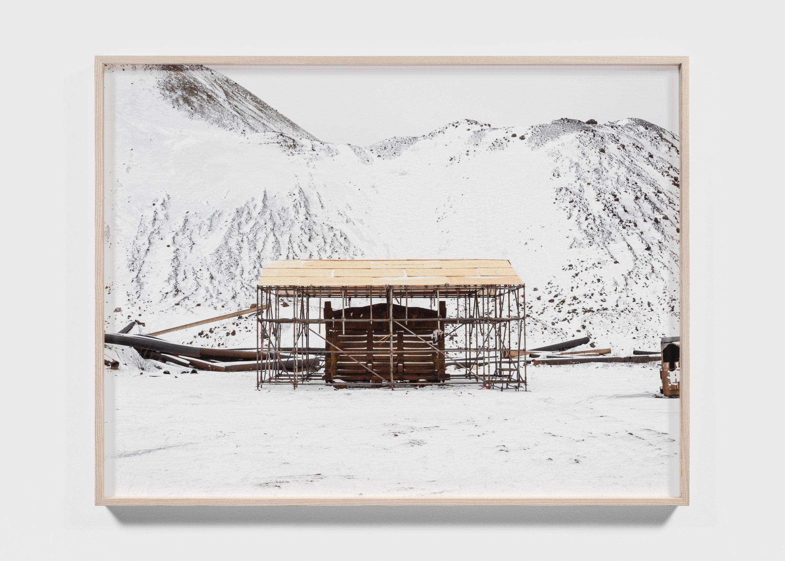 A snow-covered landscape with a wooden and metal scaffold structure in the foreground, set against a backdrop of snow-covered hills or mountains.