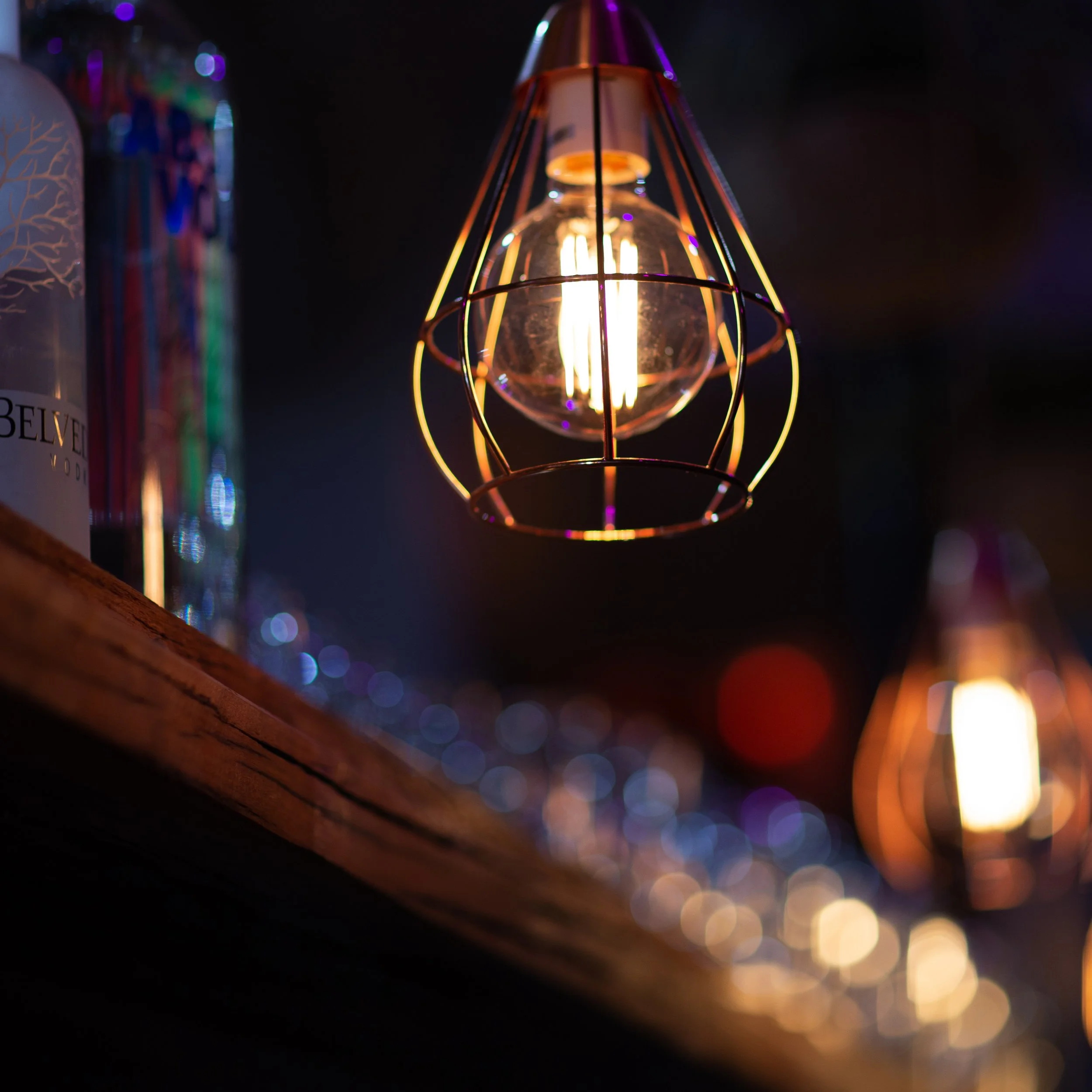 Close-up of an illuminated wireframe pendant light with blurred background of bottles on a shelf.