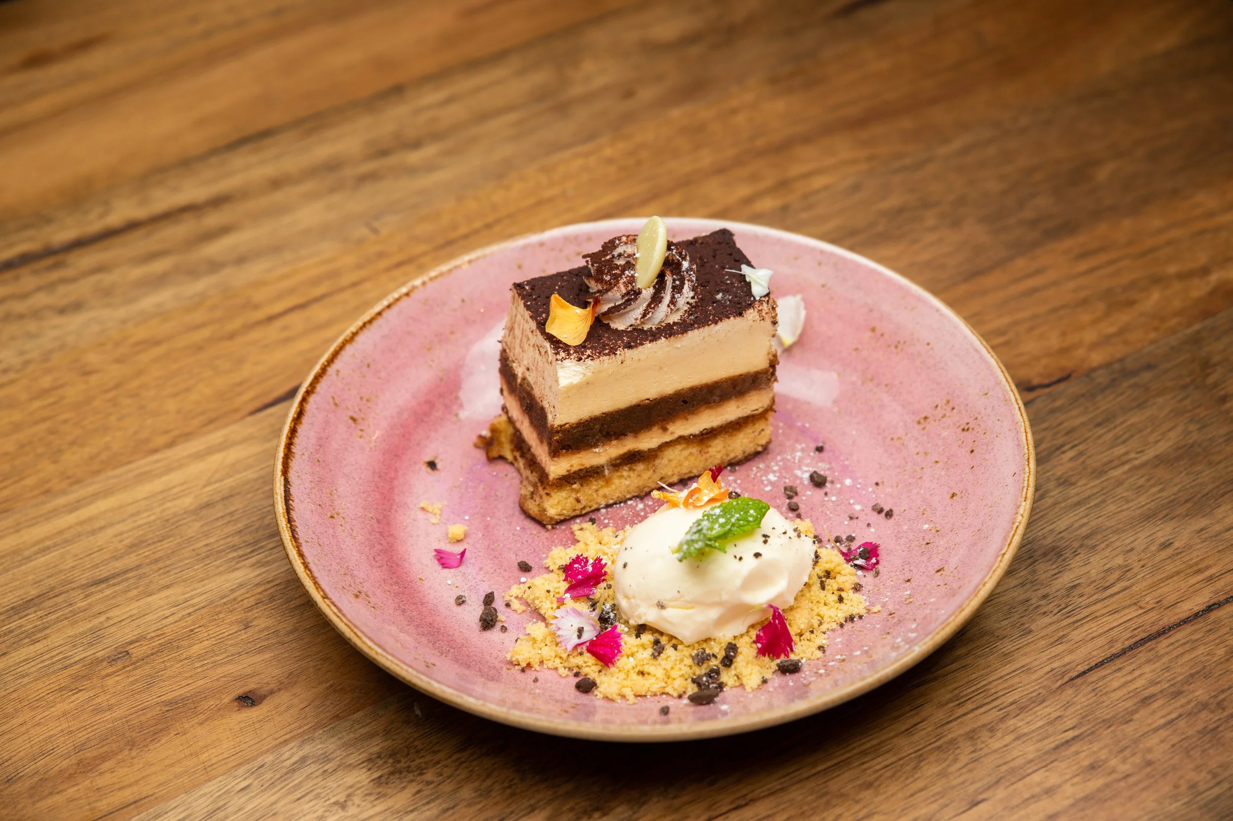 Tiramisu dessert on a pink plate with a scoop of cream, garnished with mint and edible flowers.