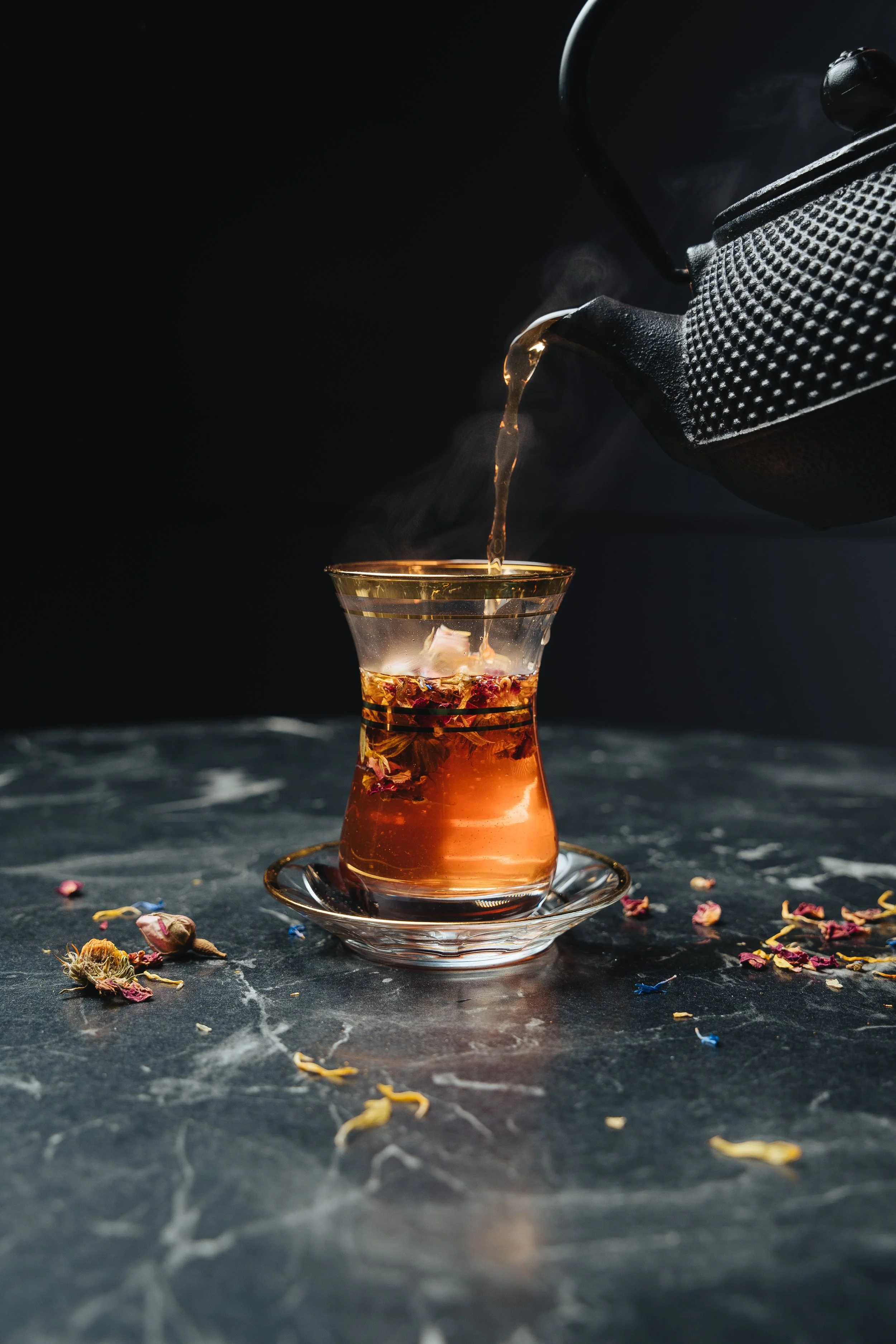 Black teapot pouring herbal tea into a glass cup on a dark marble surface, with scattered dried flowers.