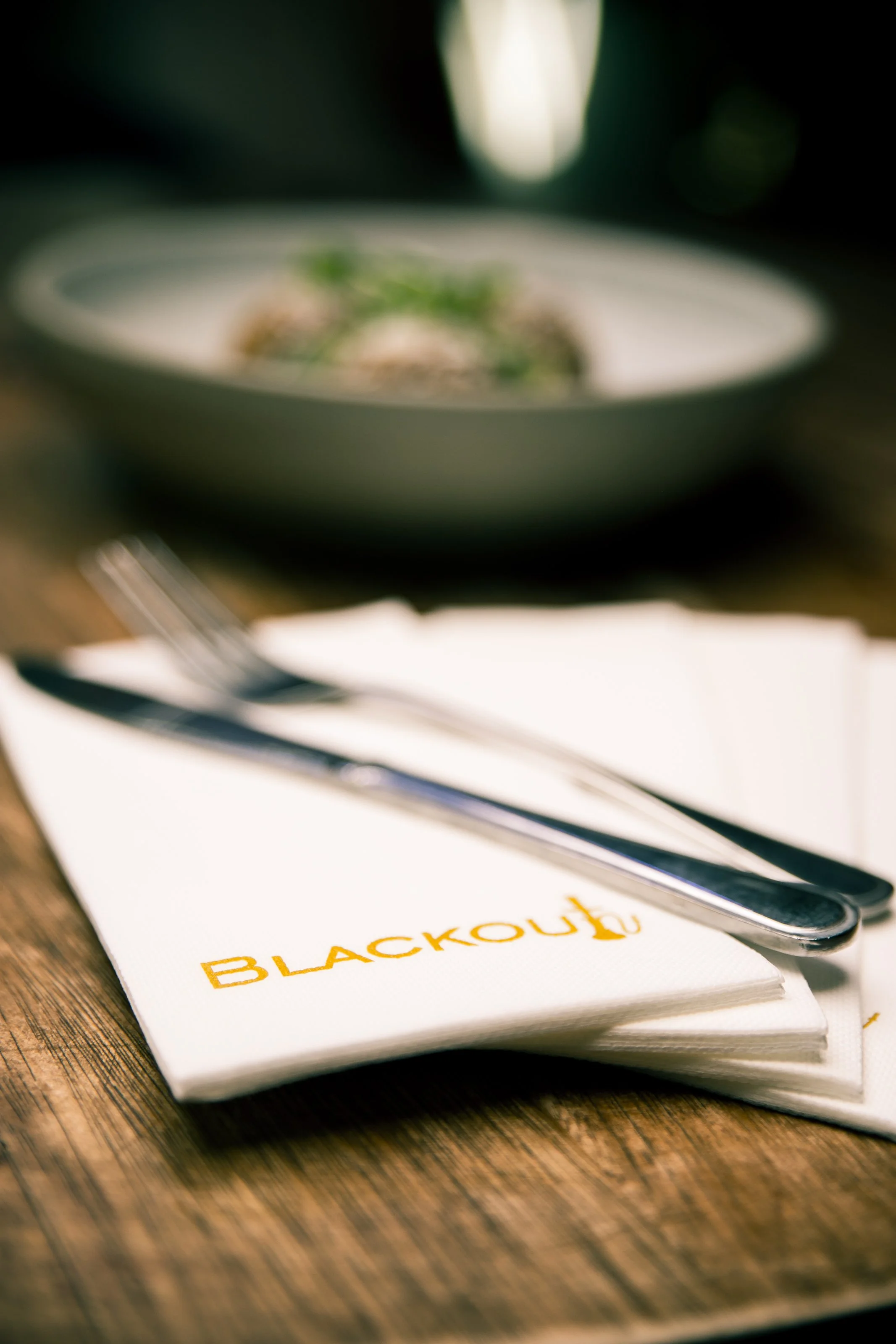 Close-up of silverware on napkins with "BLACKO" logo, blurred background of food in a bowl on wooden table.