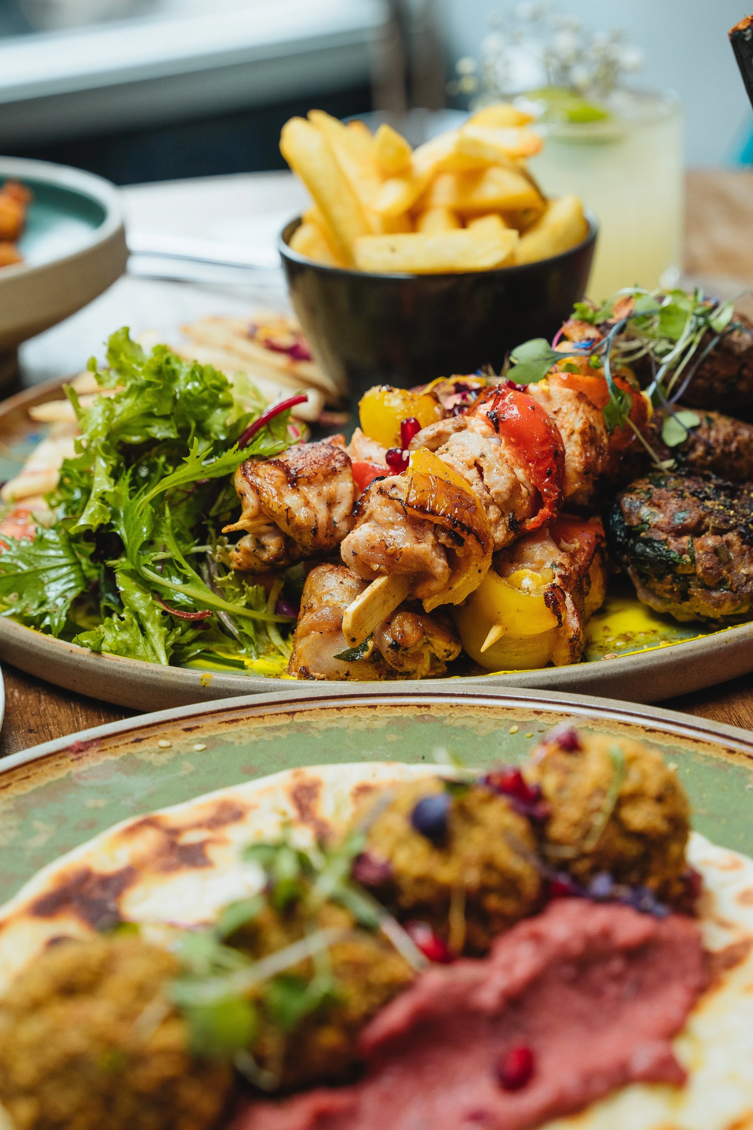 Close-up of a platter with grilled chicken skewers, fresh salad greens, and a bowl of fries, with vegetables and falafel garnished with sauce in the foreground.