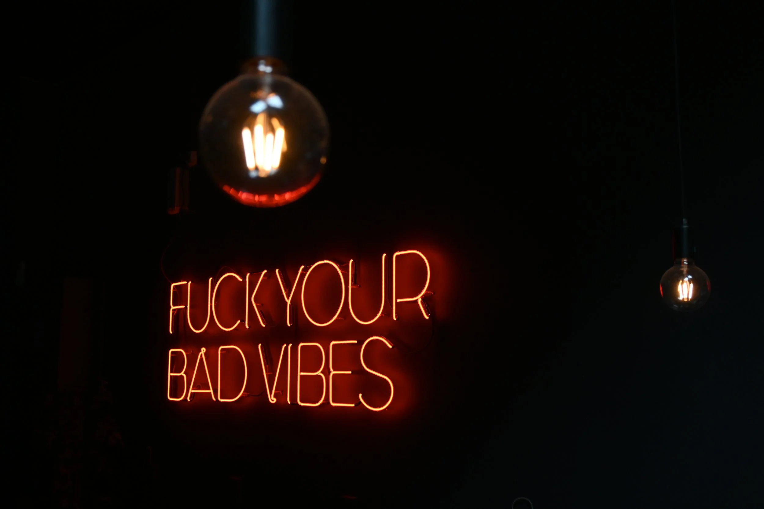 Neon sign with the words "F**k your bad vibes" against a dark background, alongside hanging light bulbs.