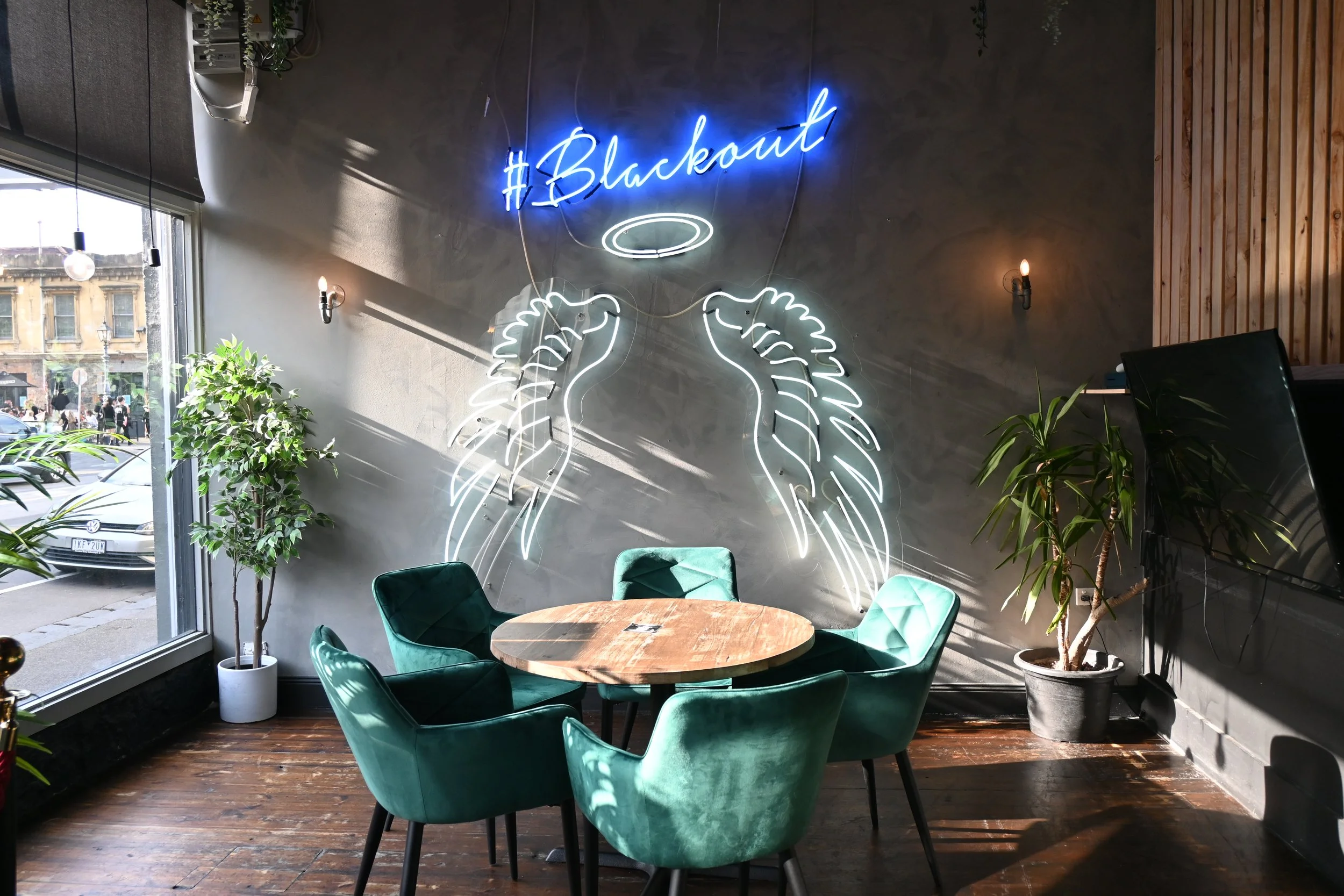 Cafe interior with green velvet chairs, a wooden round table, and neon wall art of angel wings and a halo with the hashtag 'Blackout', set against dark walls with potted plants and large windows.