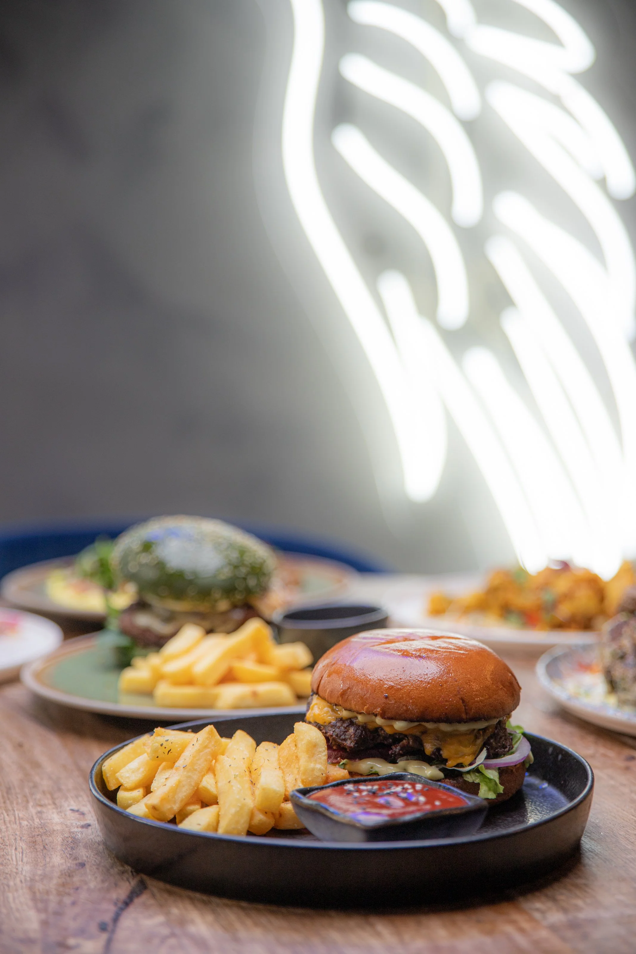 Burger with cheese, lettuce, and onion on a bun served with French fries and a side of dipping sauce on a black plate, with more food in the background on a wooden table.
