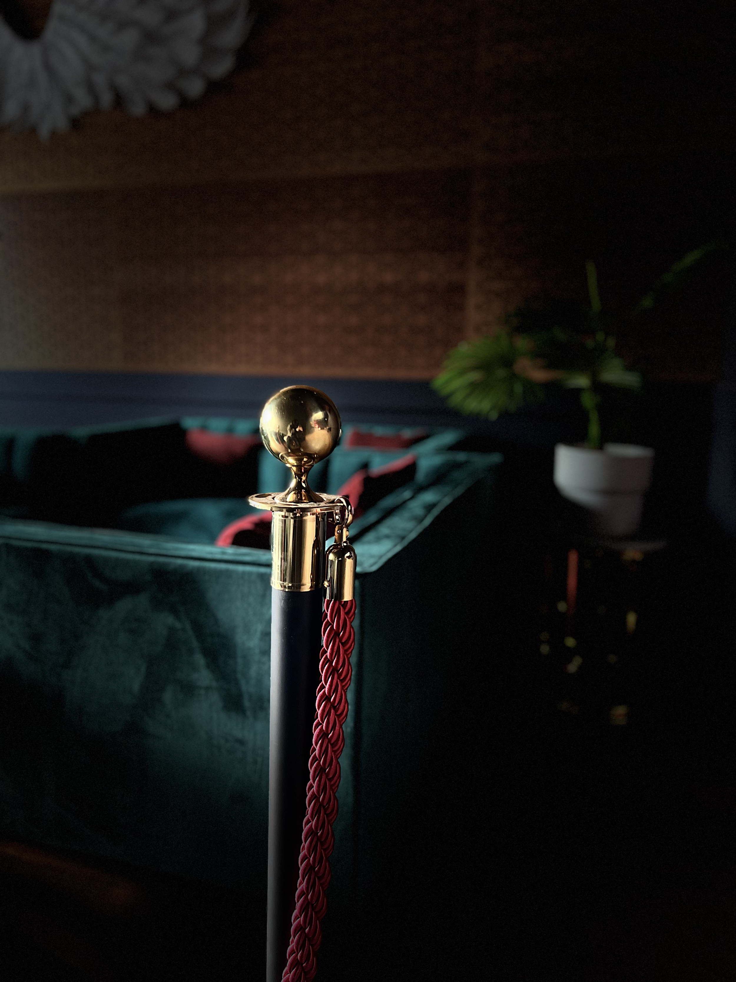 A gold stanchion post with a red velvet rope in a dimly lit room, featuring green velvet seating and a potted plant in the background.