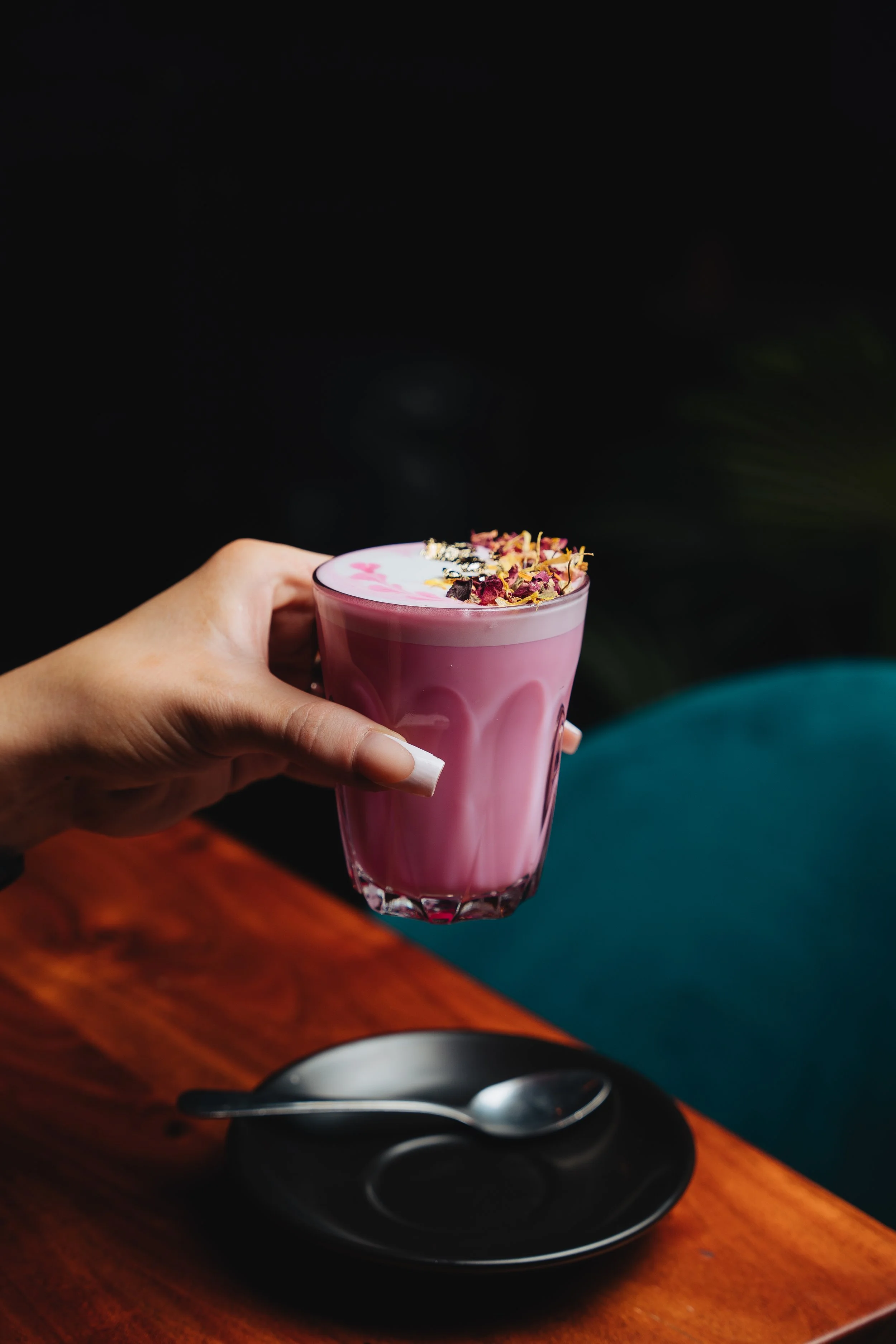 A hand holding a glass of pink latte with floral garnish, placed above a wooden table with a black saucer and a spoon.