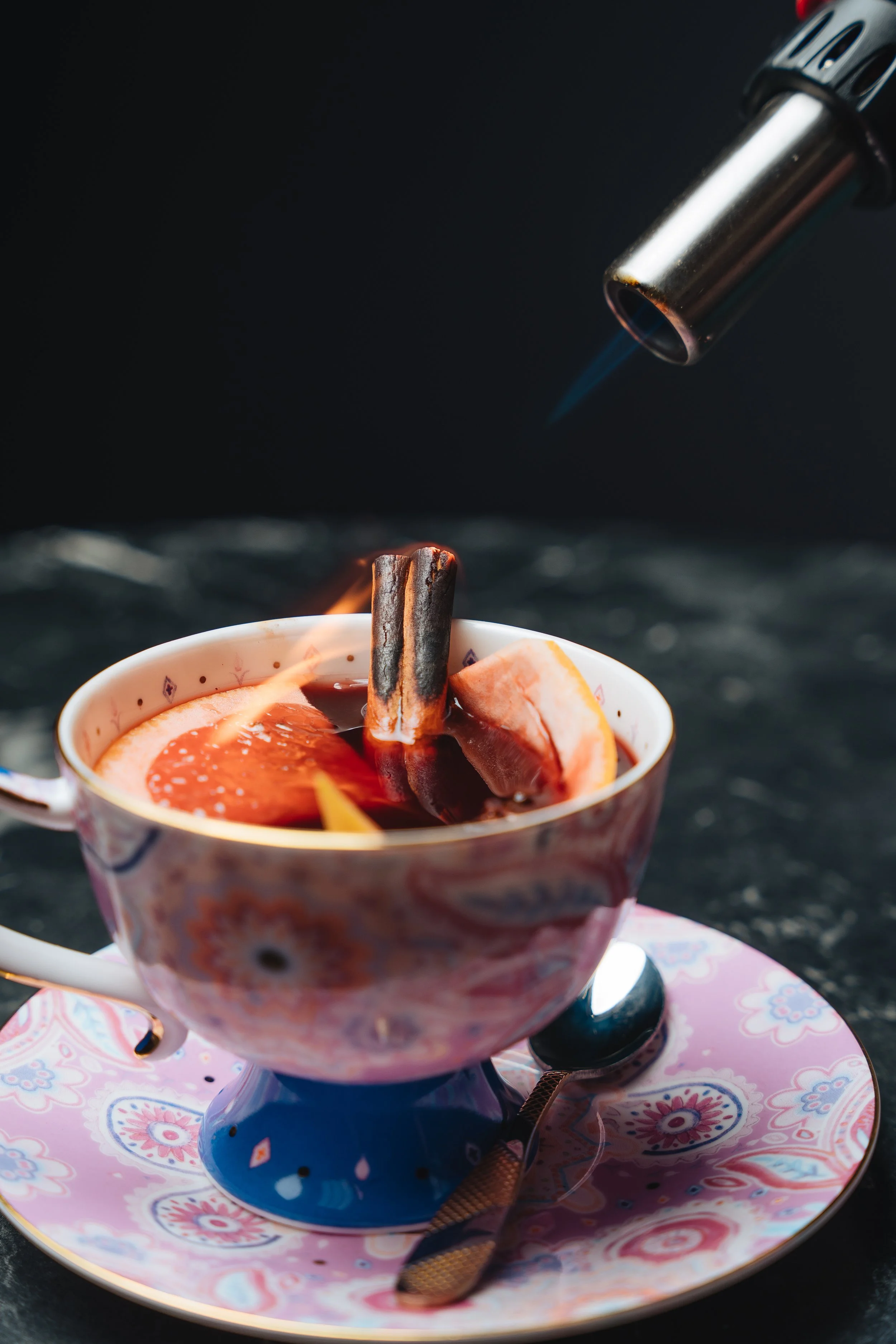 A decorative tea cup with a warm beverage, containing a cinnamon stick and citrus slices, being heated with a kitchen torch.