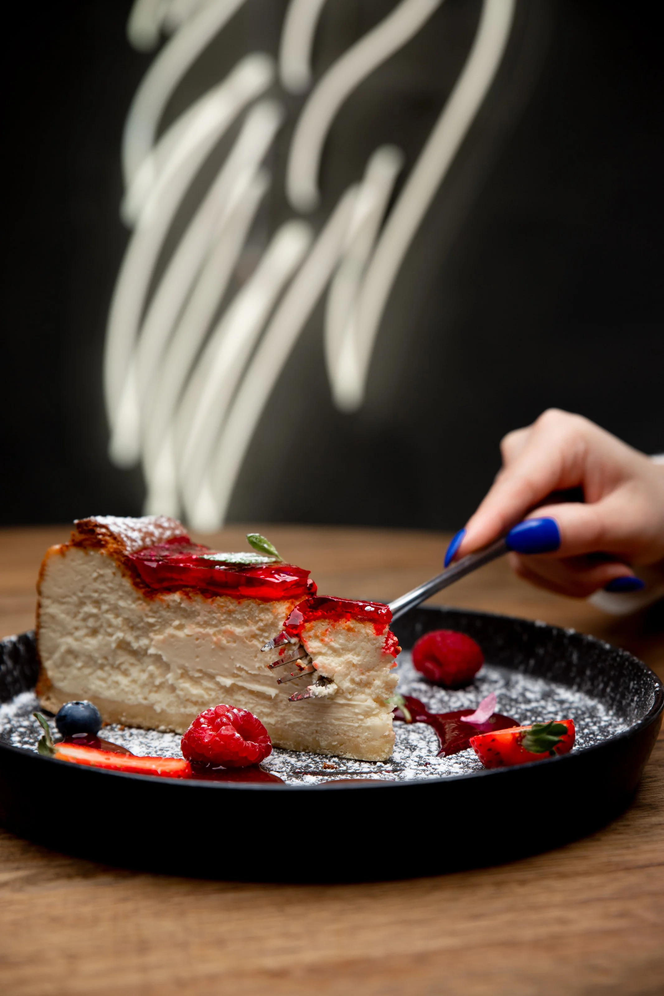Slice of cheesecake with strawberry topping and assorted berries on a black plate.