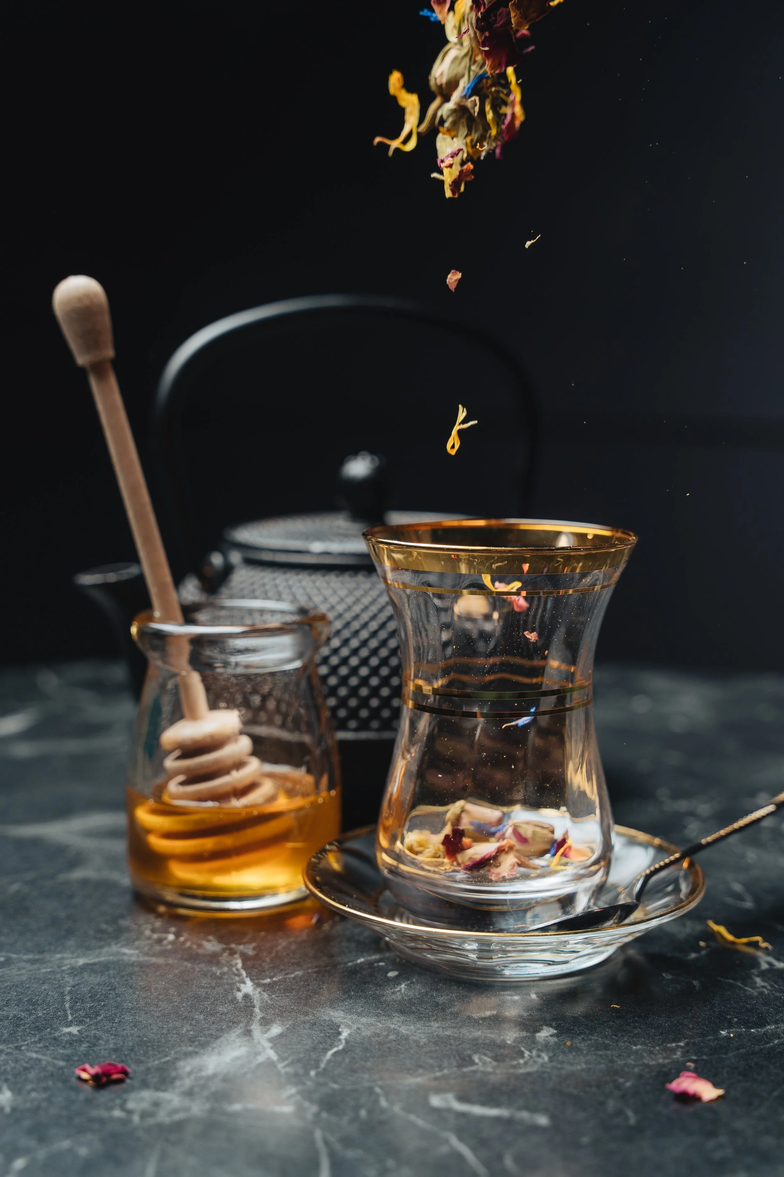 Glass jar with honey and dipper next to a clear glass with floral tea leaves, black teapot in background, on dark marble surface.