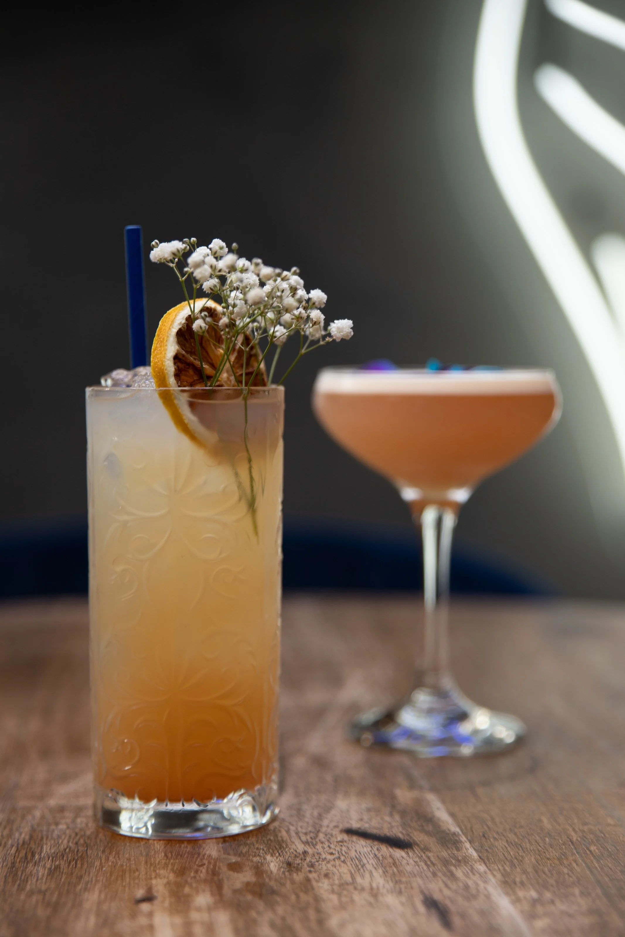 Two cocktails on a wooden table; one in a highball glass with dried citrus and flowers, the other in a coupe glass, both with garnishes.
