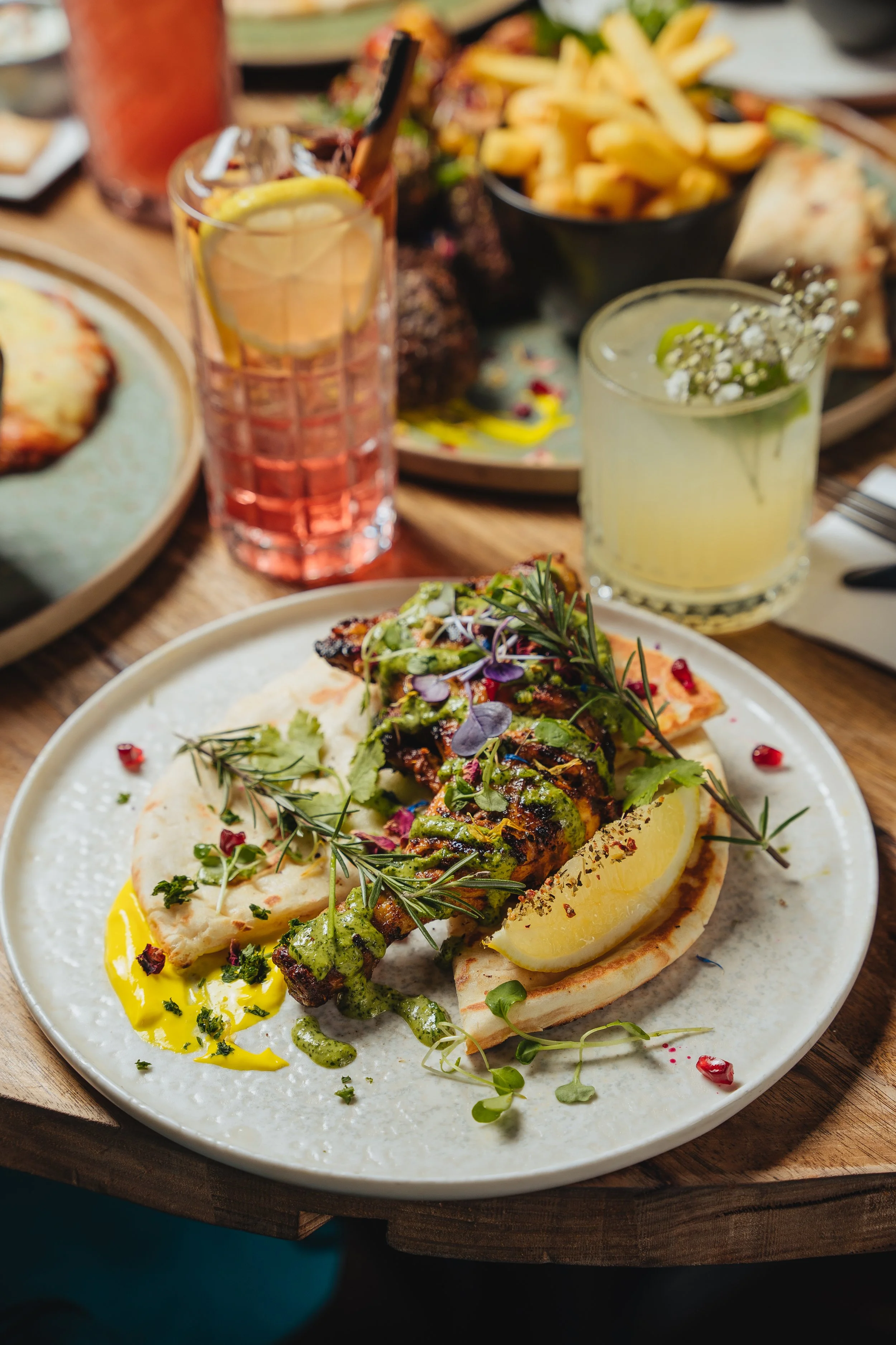 Plate with grilled chicken on flatbread, garnished with herbs and sauce, accompanied by lemon slice. Two refreshing drinks and a bowl of fries in the background.