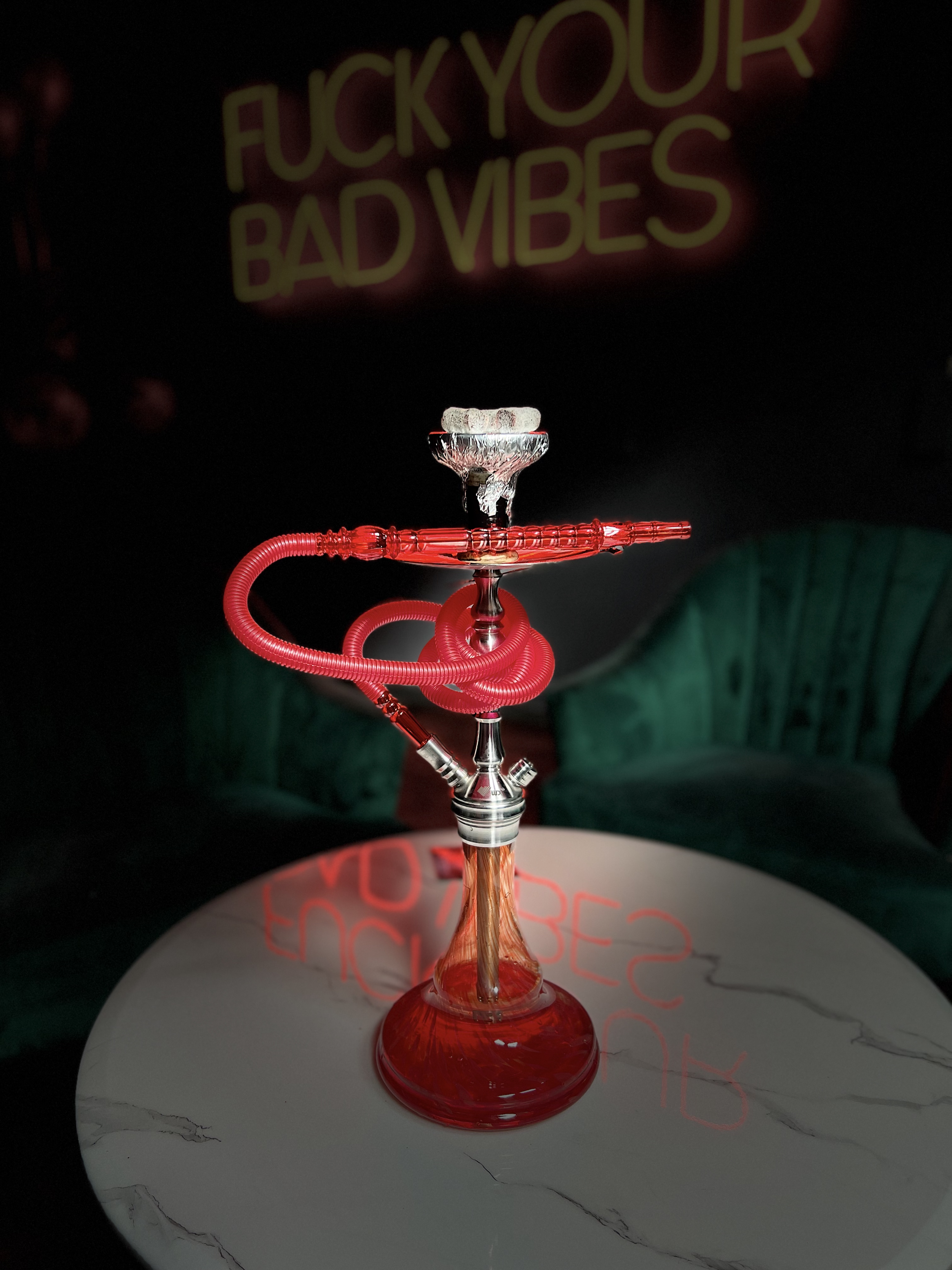 A red hookah with a matching hose on a round white marble table with a neon sign in the background.