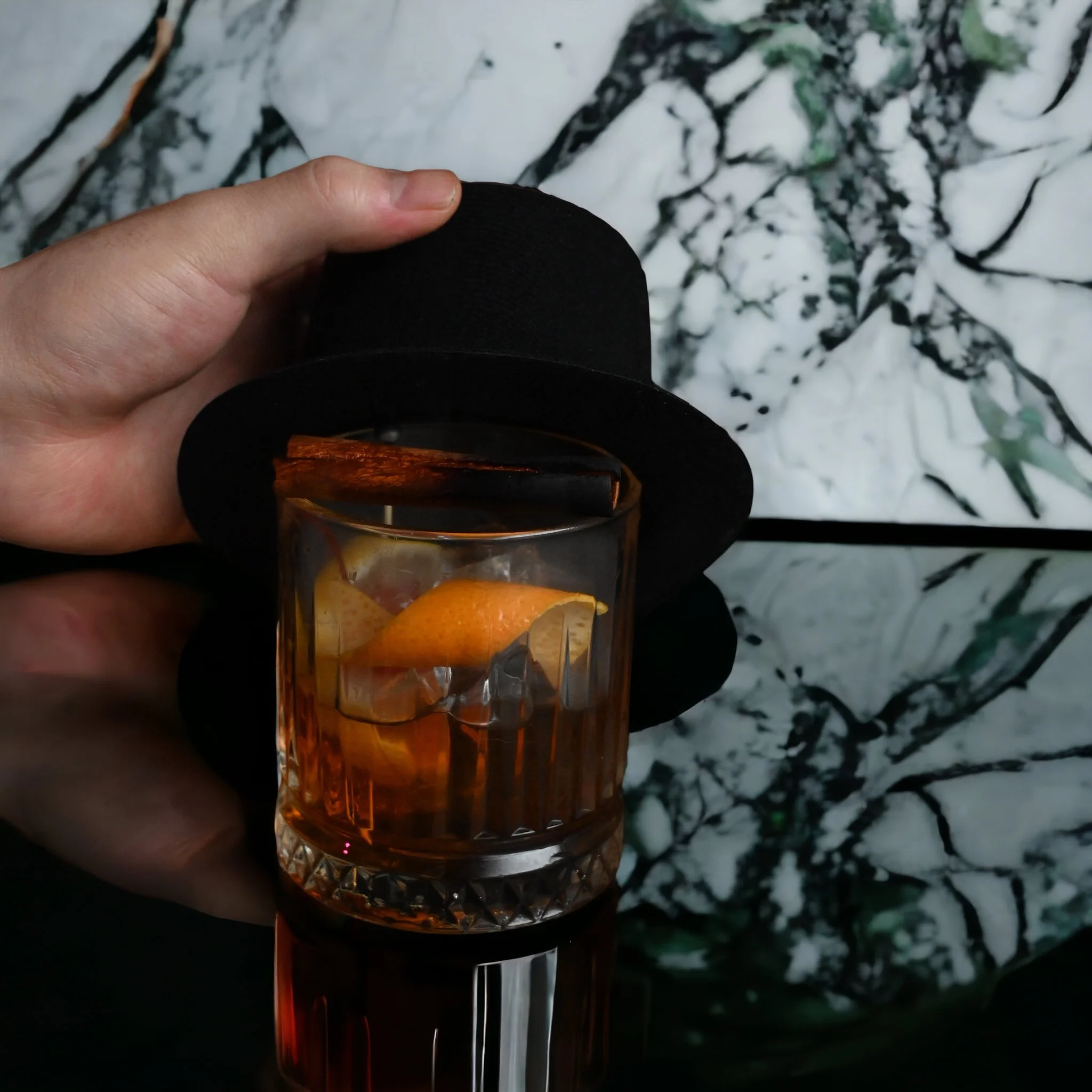 A hand holding a small black hat over a glass of whiskey with ice, an orange peel, and a cinnamon stick, against a marble-patterned background.