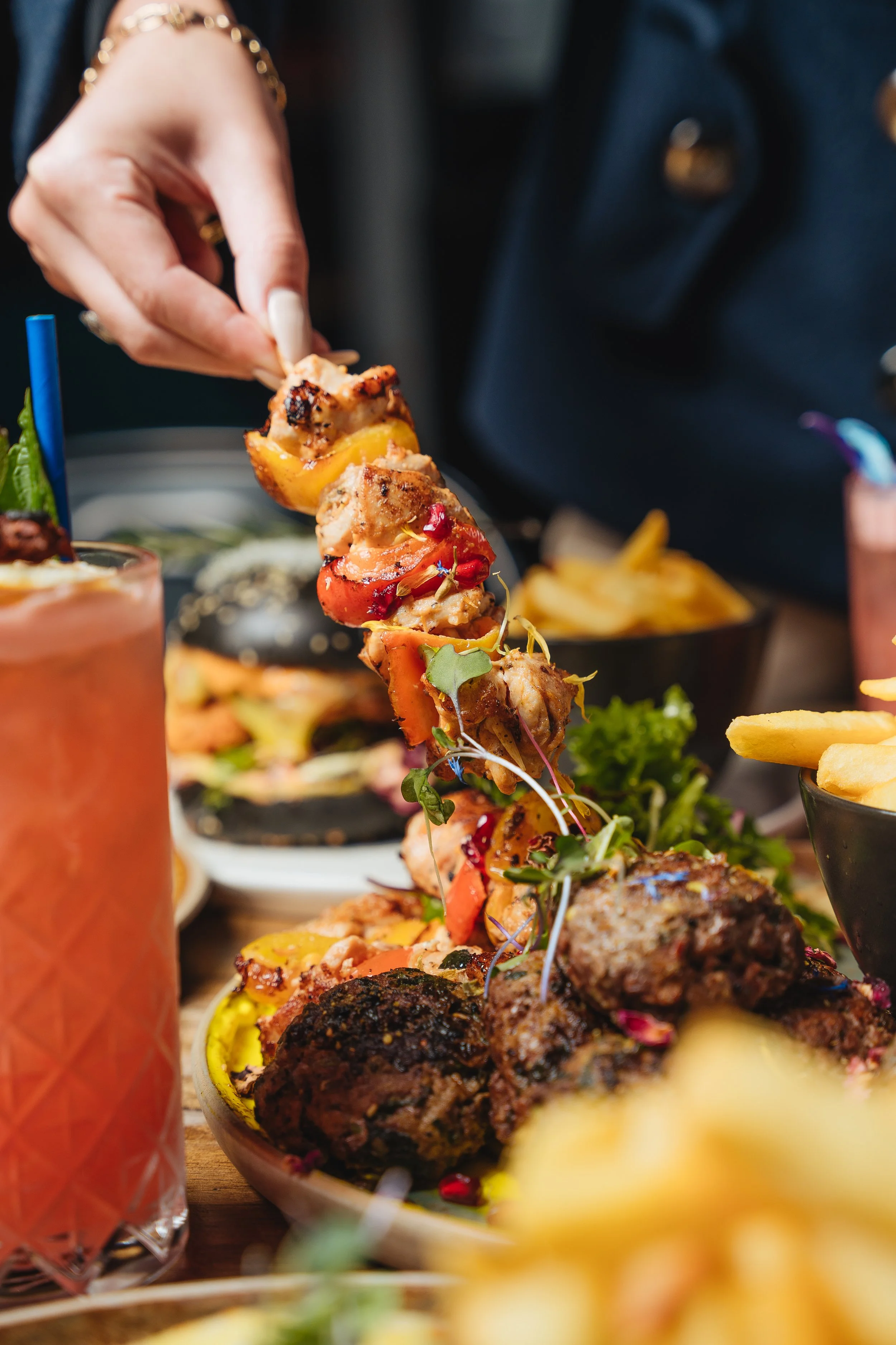 Close-up of a person holding a skewer with grilled meat and vegetables; a pink drink with a blue straw and a plate of various foods including fries are visible in the background.