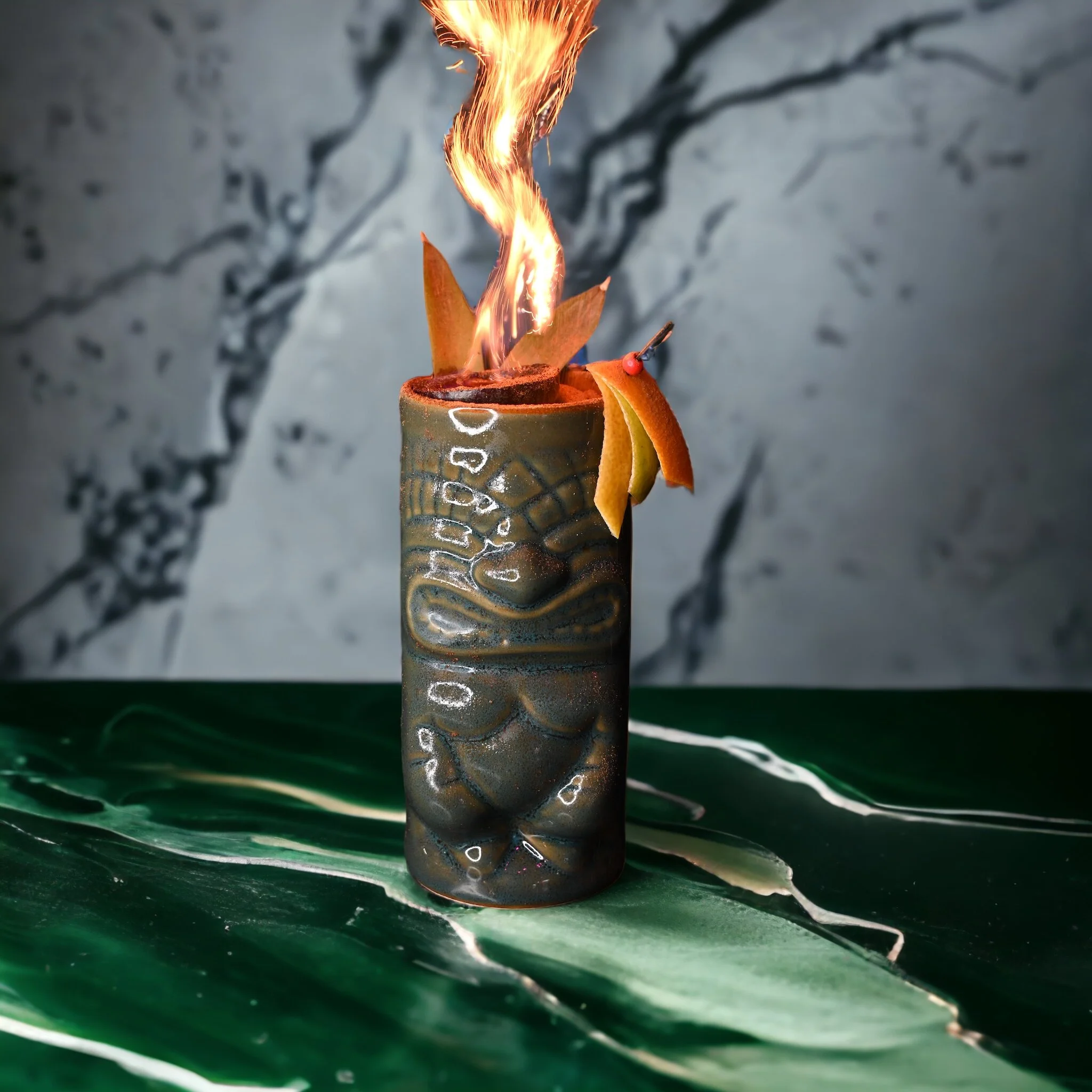 Ceramic tiki mug with flaming garnish and citrus slices on a green marble surface.
