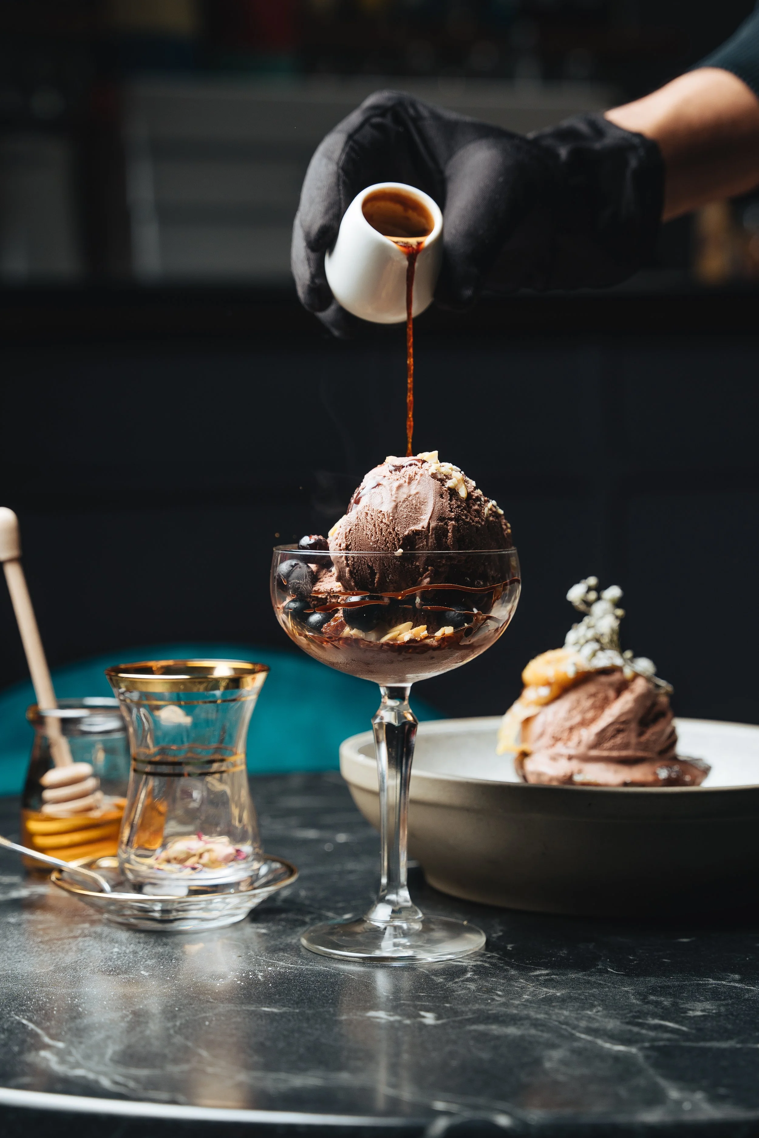 Person pouring espresso over chocolate ice cream in a glass, with honey jar and dessert plate nearby.