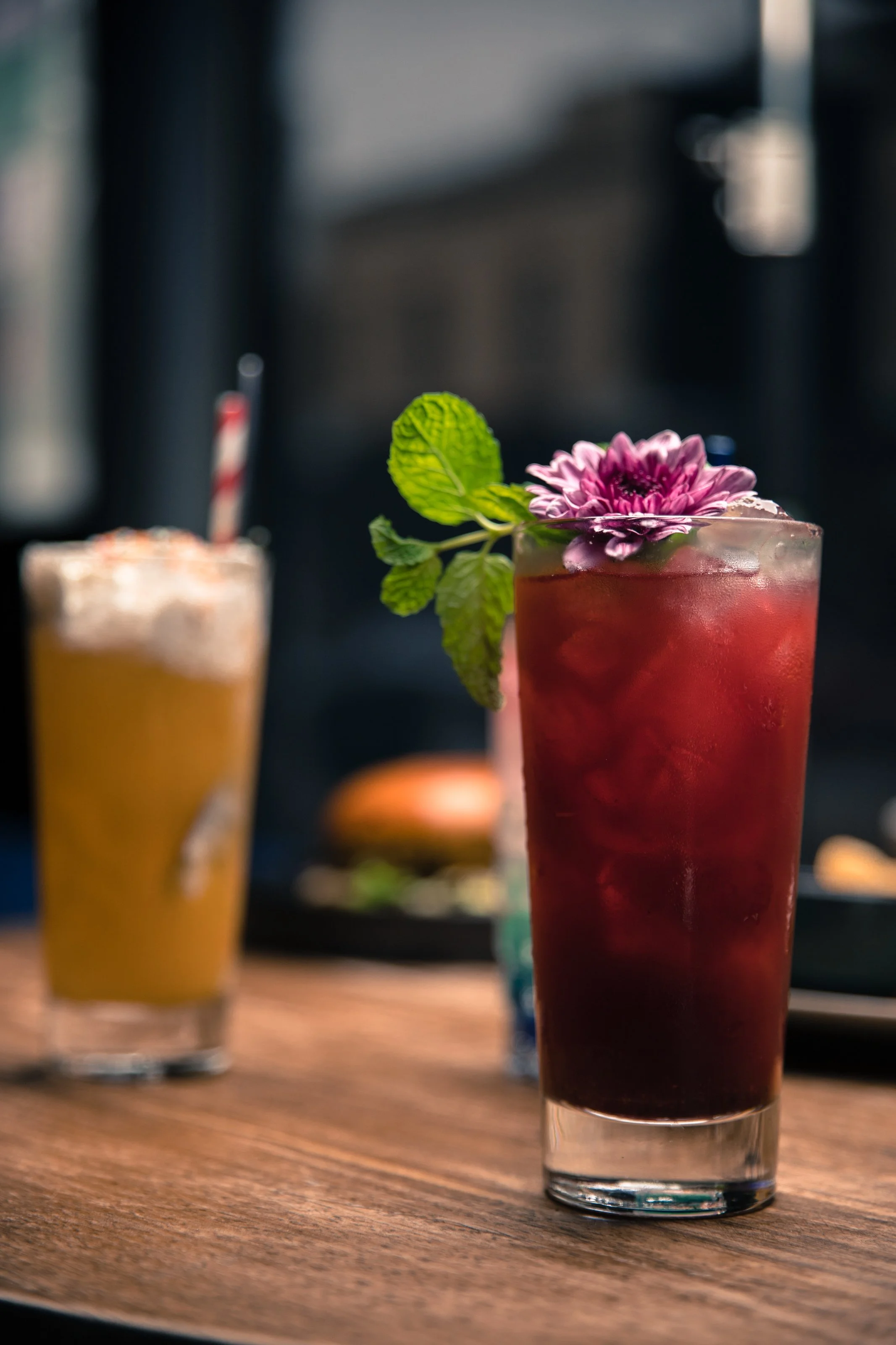Iced cocktail with flower and mint garnish on table, blurry iced drink and burger in background.