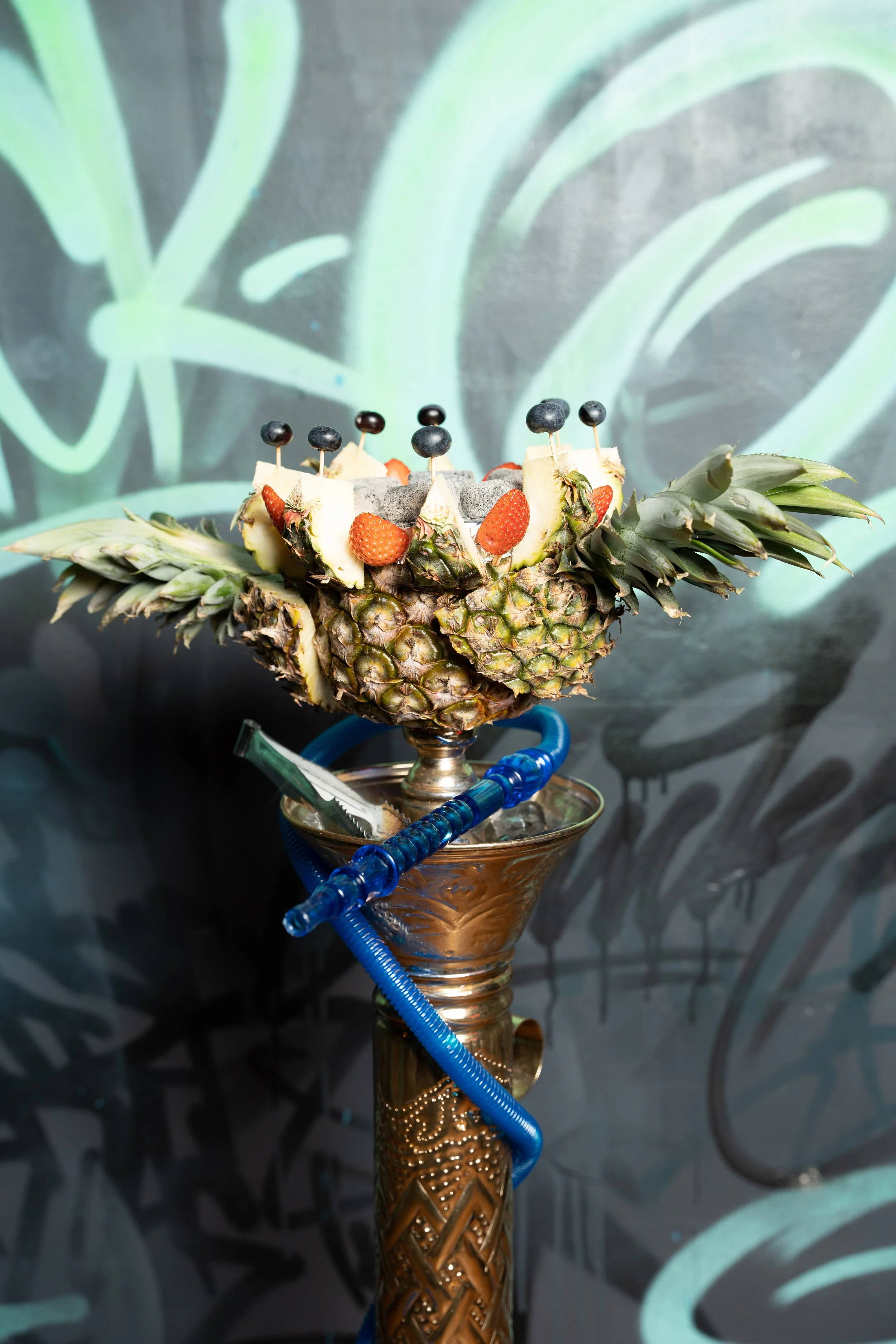A decorative hookah with a pineapple bowl top, garnished with strawberries and blueberries, set against a graffiti-style background.