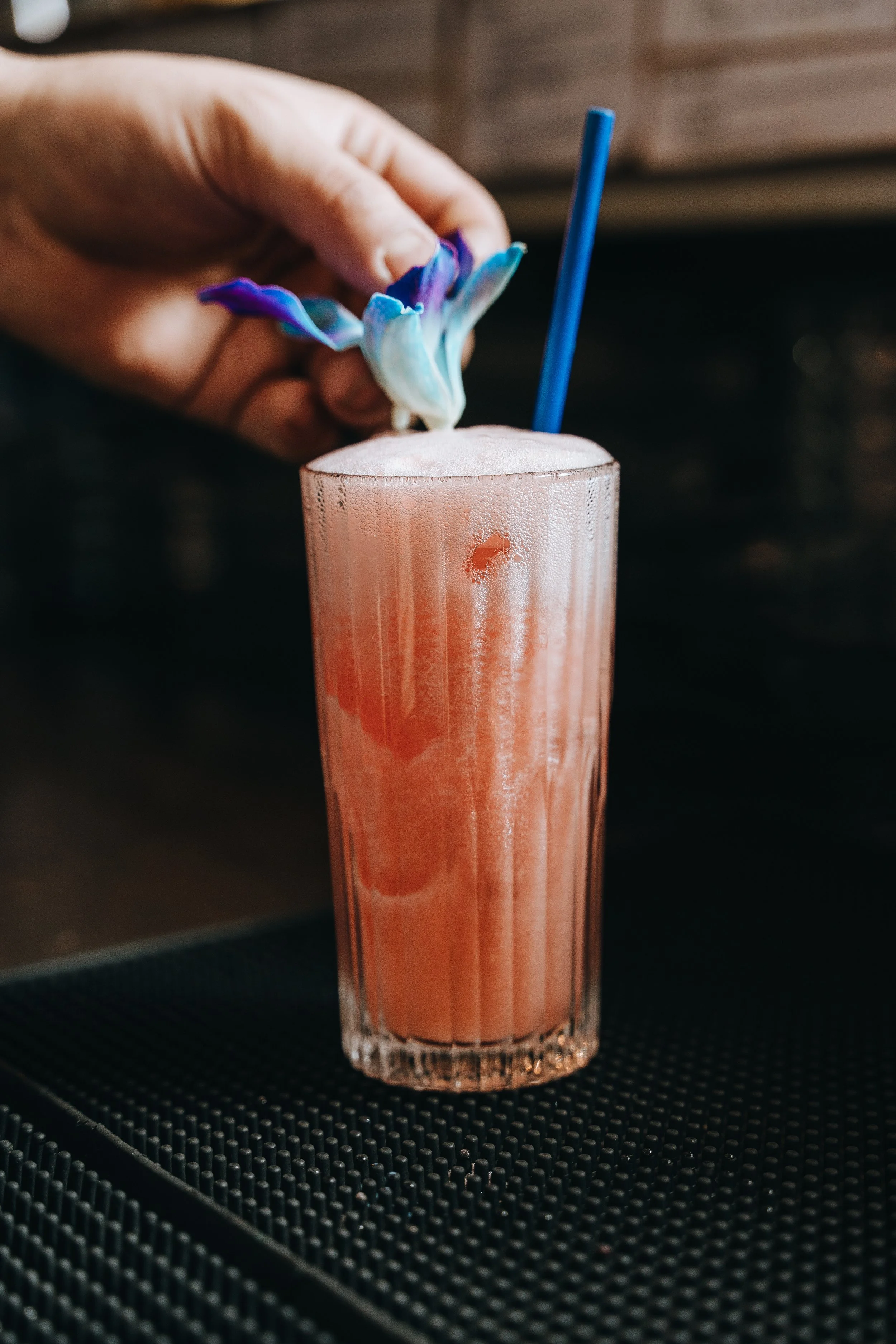 Hand placing flower garnish on pink cocktail with straw in glass.