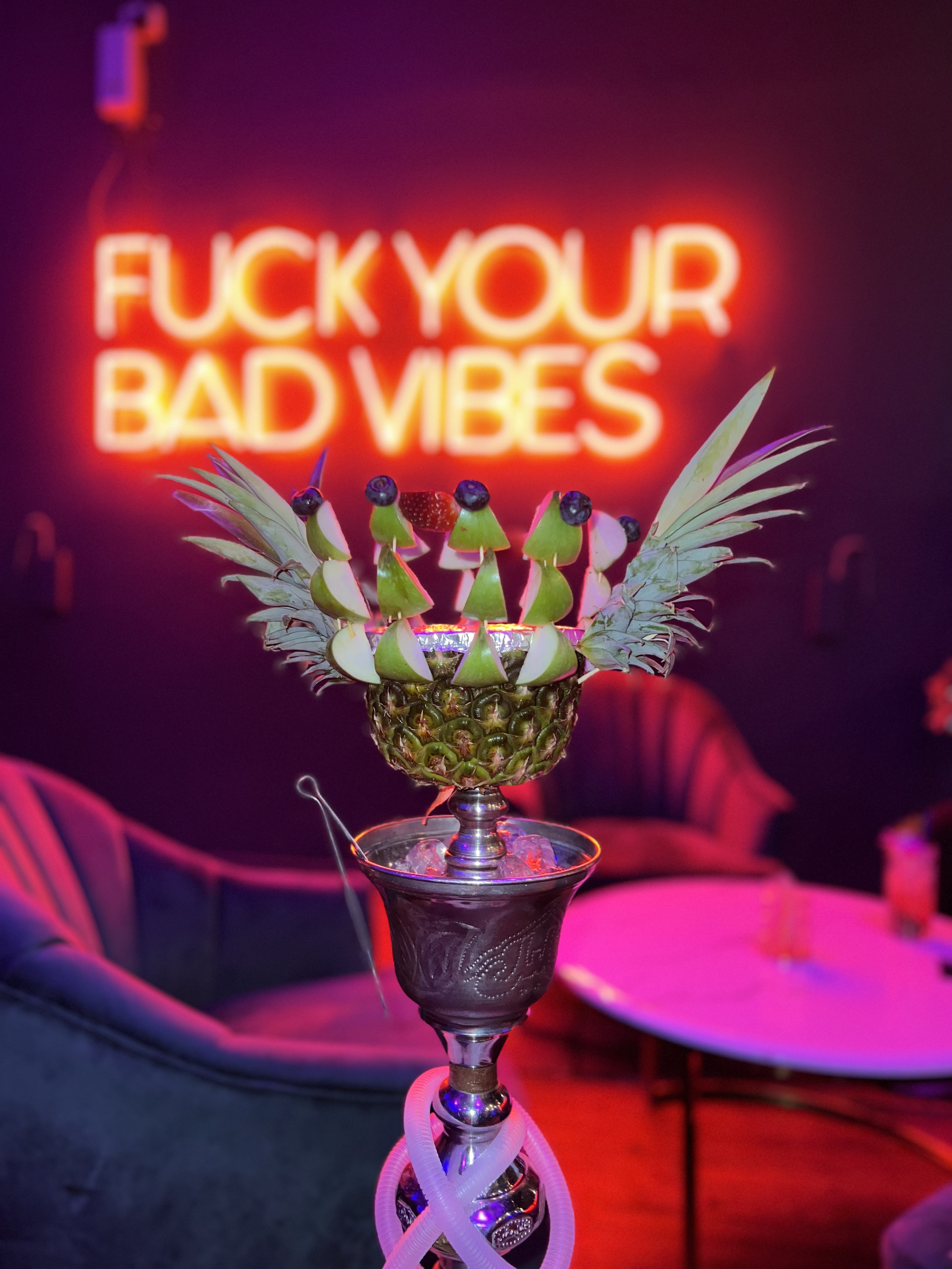 Ornate hookah with pineapple bowl and fruit garnish in dimly lit lounge, neon sign reading "F*** YOUR BAD VIBES" on wall.