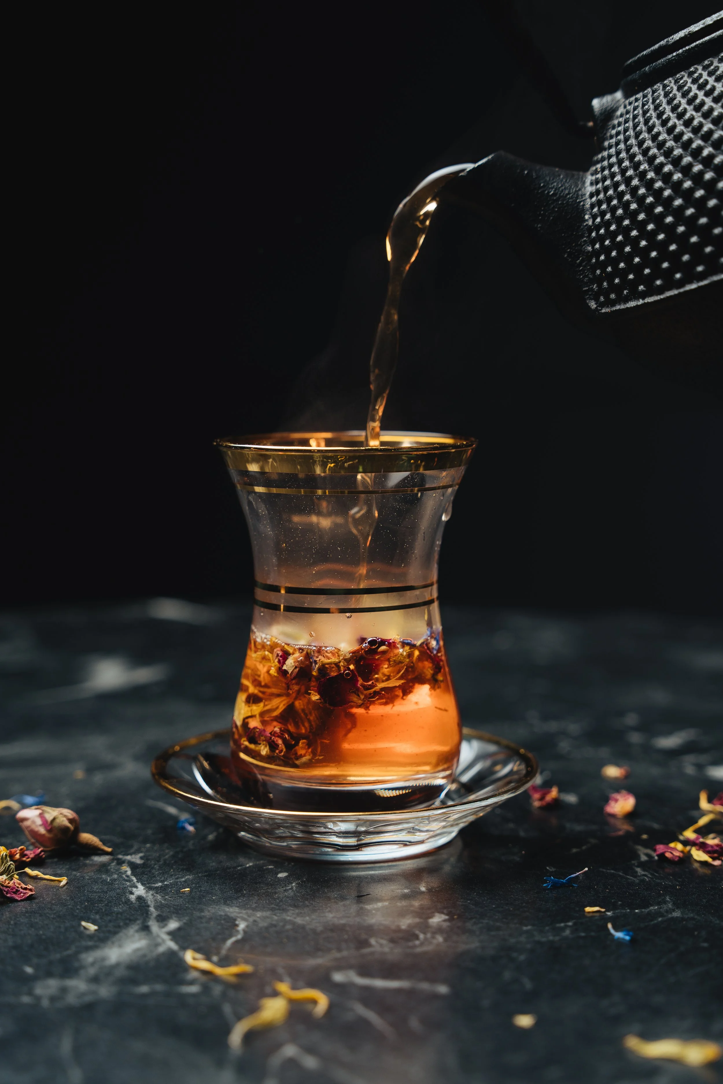 Glass of herbal tea being poured from a black teapot, with dried flowers in the glass, on a marble surface.