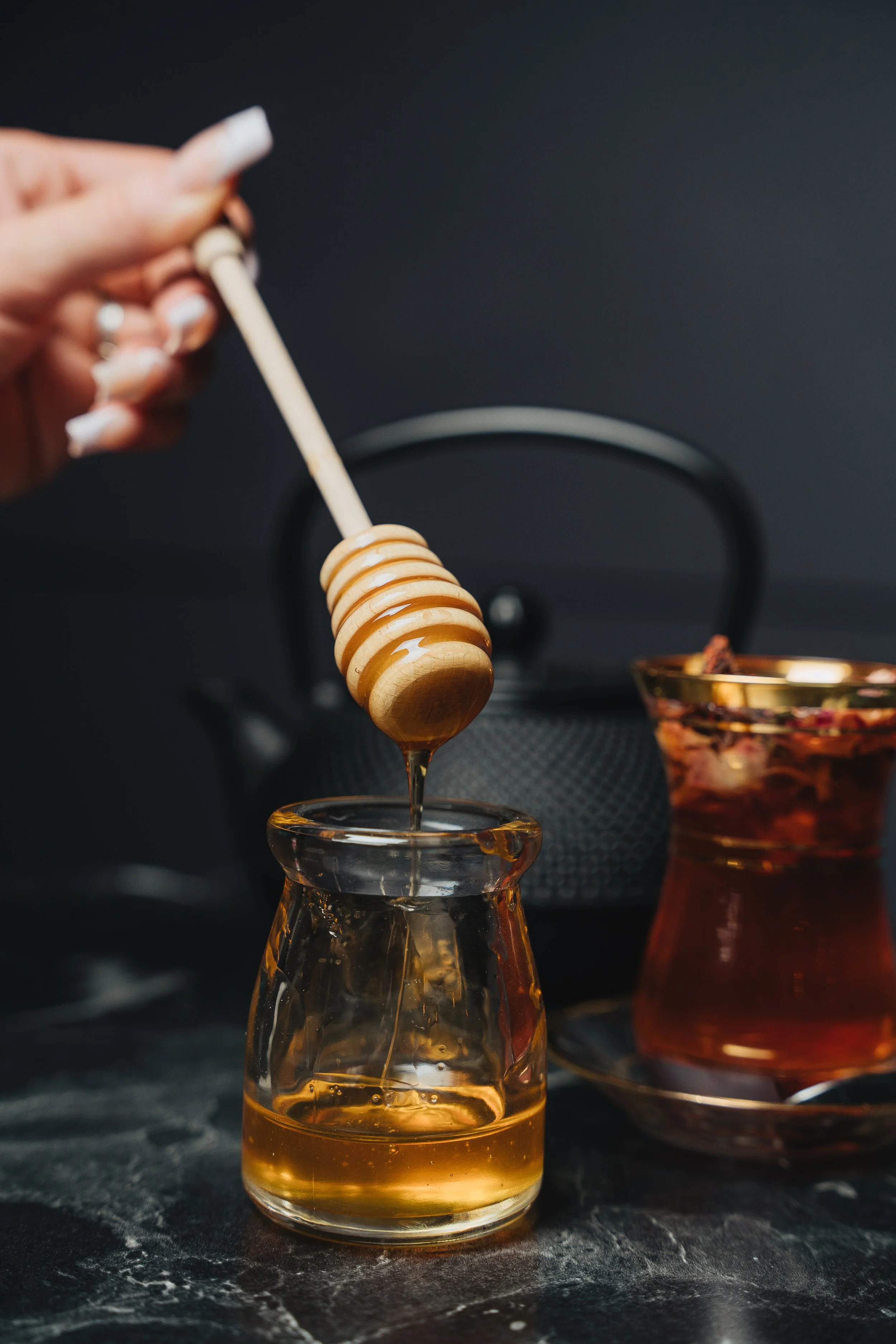 Honey being drizzled from a dipper into a glass jar, with a teapot and glass of tea in the background.