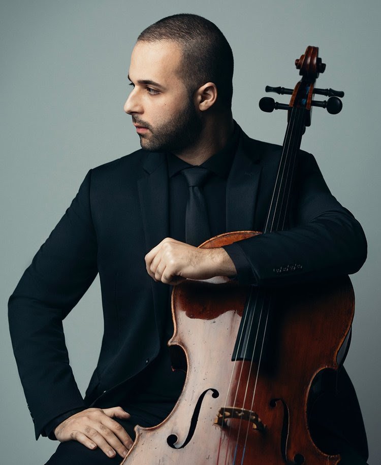 A man in a black suit holding a cello, with a serious expression, against a plain gray background.