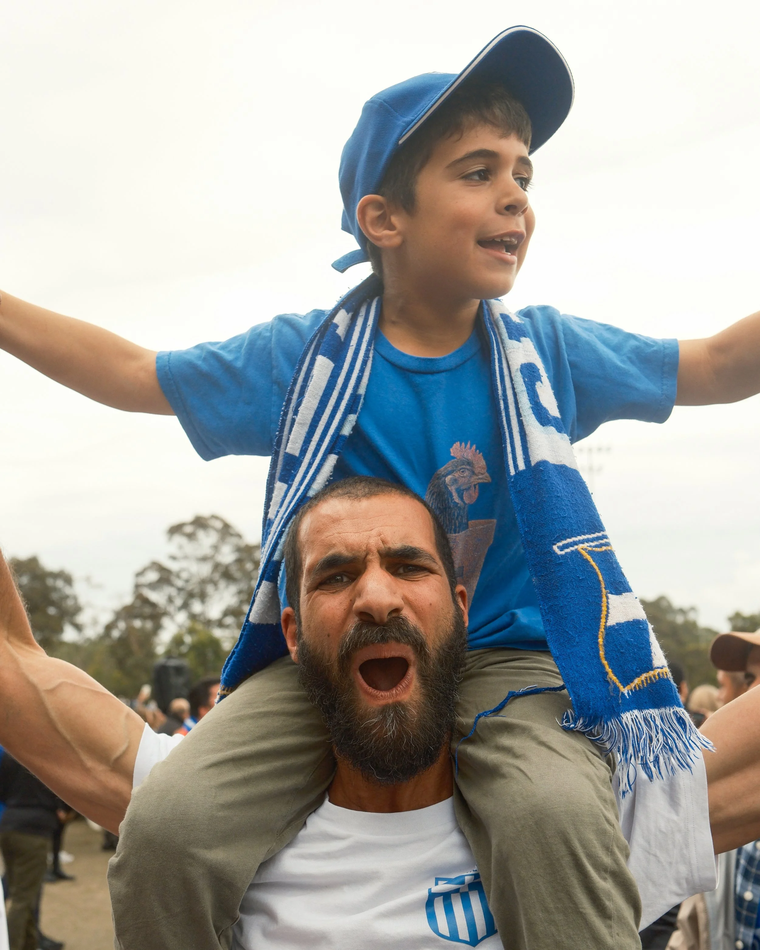 A man with a beard and a white T-shirt with blue text, carrying a boy on his shoulders during an outdoor gathering. The boy is wearing a blue cap, a t-shirt with a rooster graphic, and a blue and white scarf. The boy has his arms extended outward, sm